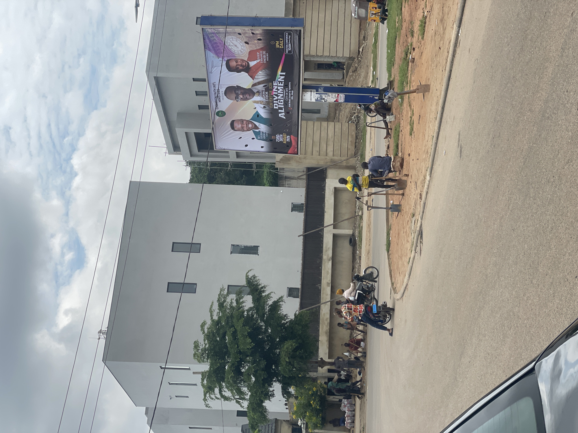 The image depicts a street scene in Guzape, Nigeria, characterized by modern architecture and a billboard advertising a religious event. In the background, two contemporary buildings with minimalist design dominate the skyline. A large billboard, featuring portraits of three men and the text "DIVINE ALIGNMENT" and "CHAMPION FAITH INTL MINISTRIES," is prominently displayed.
In the foreground, a road curves through the scene. Several individuals are present, some gathered near the billboard and others on motorcycles. One man is seated on a rock while others stand around him, holding shovels, suggesting some form of construction or maintenance work. Two motorcycles are visible on the road, one with a rider and a passenger, and another with a single rider. Various bags of materials are stacked on the side of the road. The overall environment appears to be an urban setting with evidence of ongoing development and daily life. The sky is overcast with scattered clouds.