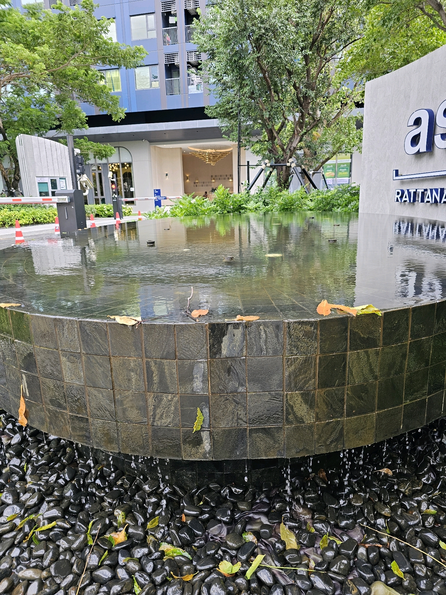 The image captures a contemporary water feature located outside a building in the city of Nonthaburi City Municipality, Thailand. The water feature is a circular fountain with dark, tiled sides, surrounded by black, polished pebbles. Water gently spills over the edge of the fountain, creating a shimmering effect on the pebbles below. Fallen leaves, both green and brown, are scattered on the fountain's surface and among the pebbles, suggesting an outdoor setting with natural elements.
In the background, a modern building with blue and gray facades dominates the scene. The building features multiple levels with balconies and numerous windows. Trees with lush green foliage are interspersed around the building and the water feature, adding to the ambiance of a well-maintained urban landscape. A white stone monument with the letters "as" and "RATTANAM" visible in blue, stylized font, stands to the right of the fountain. The area appears to be an entrance or public space, with a security barrier, traffic cones, and an automatic gate visible on the left side of the frame. The lighting suggests it is daytime, with overcast conditions as indicated by the soft light and reflections on the water.