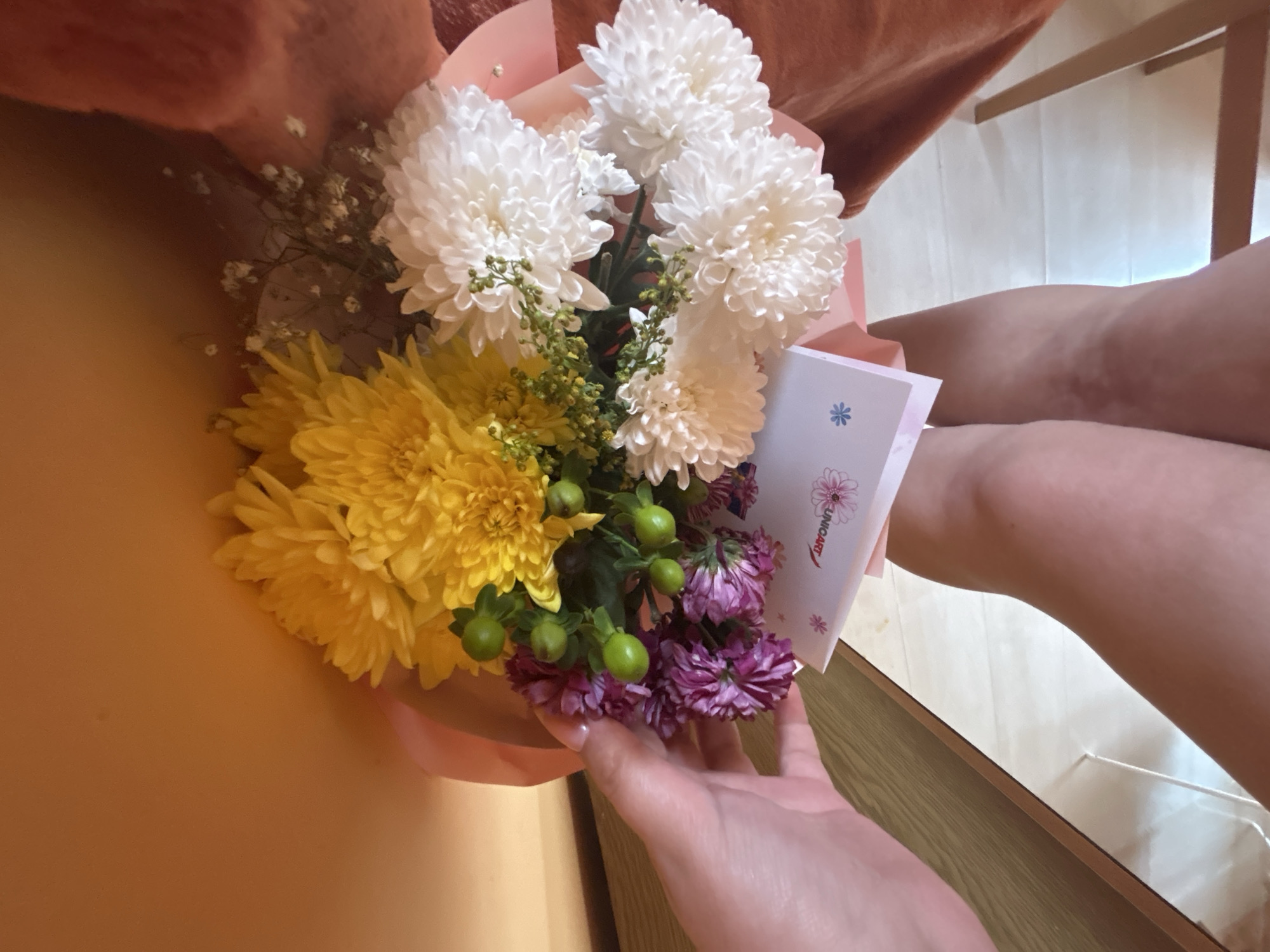 A person is holding a bouquet of flowers, composed of white chrysanthemums, yellow mums, purple asters, and other green foliage. The flowers are wrapped in a pink paper. A greeting card with the word "UNIQART" is visible within the bouquet. The person's hand and legs are visible. The scene is indoors with a wooden floor. It is impossible to determine the specific location in Blagoevgrad, Bulgaria, from this image alone.