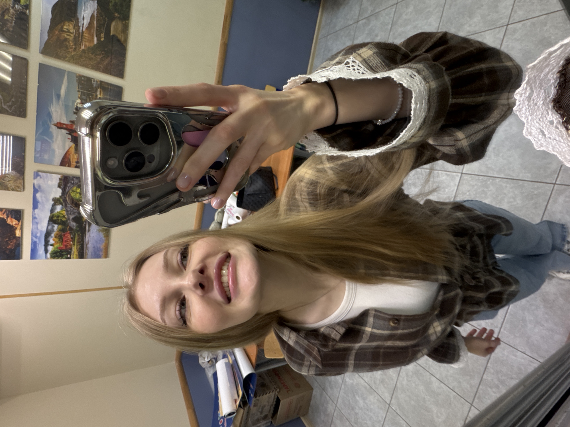 A young woman takes a selfie in what appears to be an indoor setting. She is wearing a brown plaid shirt with lace accents, a white t-shirt underneath, and blue jeans. She is holding a smartphone with a silver case in her left hand, and the camera is pointed at her face. Her blonde hair is long and flowing. In the background, there are tiled walls, a row of landscape pictures on the wall, and some furniture. A box sits at the bottom of the shot. There is no explicit evidence in the image indicating that the location is Perm, Russia.
