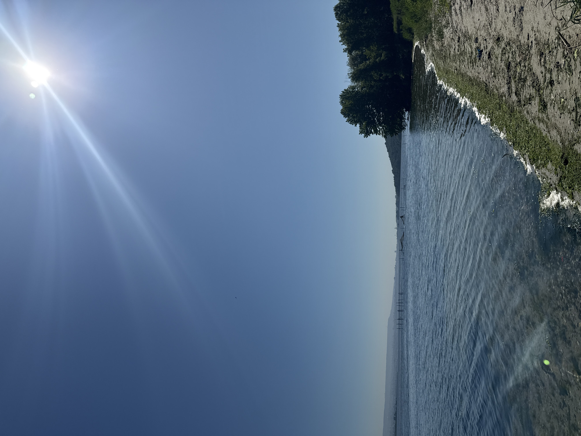 The image shows a body of water, likely a lake or river, with a shoreline on the right. The shoreline transitions from water to a sandy or gravelly area with sparse green vegetation. A line of trees runs along the shoreline further back. In the distance, the opposite shore of the water body is visible, appearing hazy. The sky is a gradient of blue, with the sun shining brightly in the upper left, creating lens flares. The perspective is slightly angled, giving a sense of depth to the scene. The location is Konstantinovo, Bulgaria. There are no people visible in the frame. The image does not capture any actions or interactions.