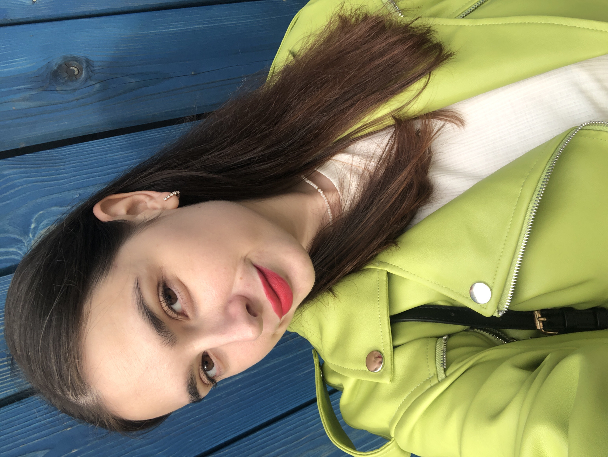 This is a vertical, close-up, eye-level selfie of a young woman with long, dark brown hair and brown eyes, wearing bright lime green faux leather jacket. She is also wearing a white top underneath and bright red lipstick. She has a delicate chain necklace made of small white beads and a small diamond stud in her left ear. The background is composed of weathered blue wooden planks. The image appears to have been taken outdoors on a bright day, indicated by the lighting and lack of shadows. The woman is looking directly at the camera with a slight smile. The setting suggests a casual outdoor environment, possibly a park bench or a deck. The overall mood of the image is cheerful and confident. No text is visible in the image.