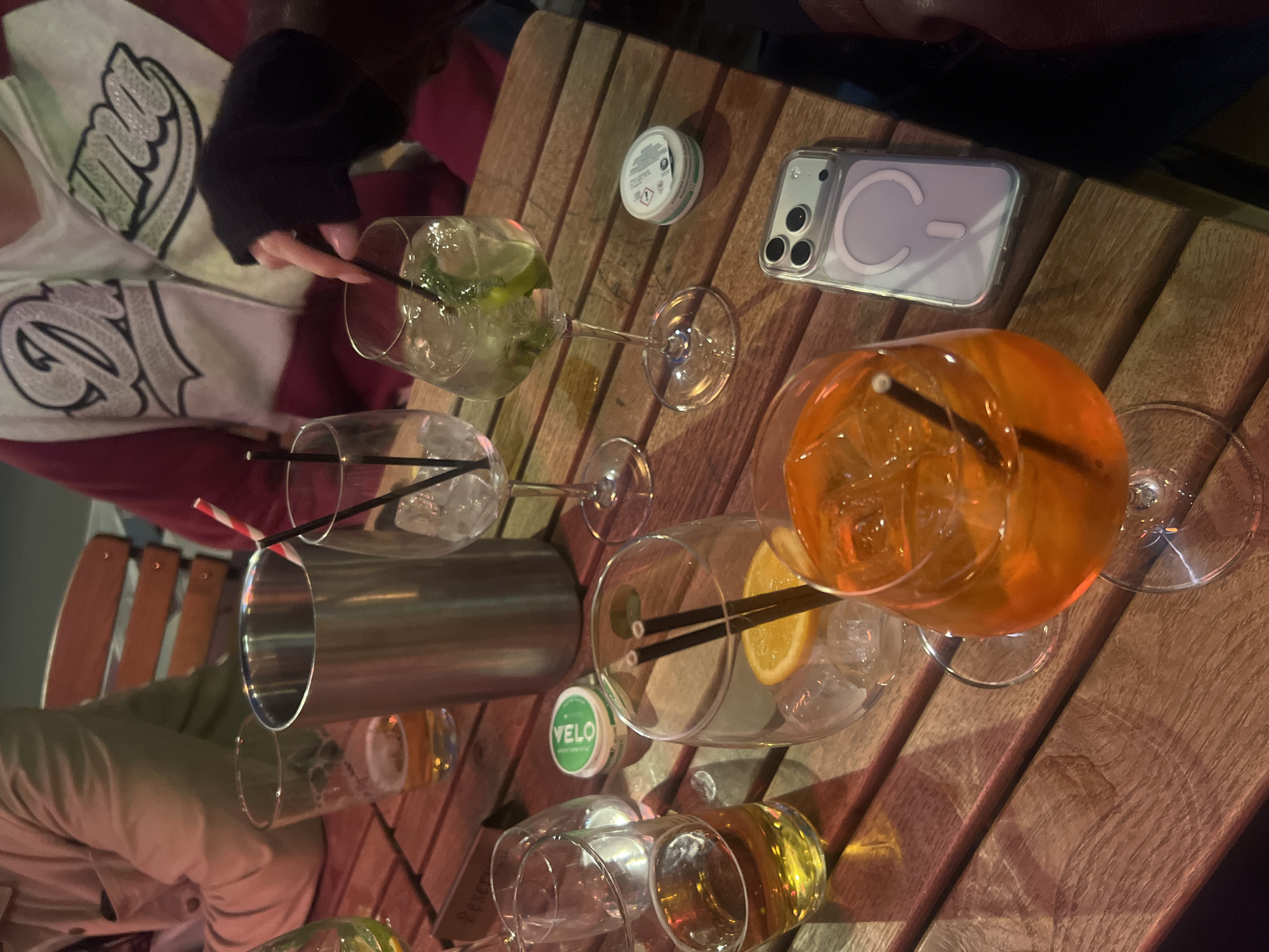 The image captures a casual gathering on a wooden table, likely at an outdoor setting in London, United Kingdom. Several glasses are present, some filled with beverages: one contains a clear liquid with lime and mint, another a vibrant orange drink with ice and an orange slice. There's also a stainless steel cocktail shaker. A white tin of Velo nicotine pouches and a white iPhone with a clear case are also on the table. Garments of people are visible around the table, suggesting a group engaging in conversation or drinks.