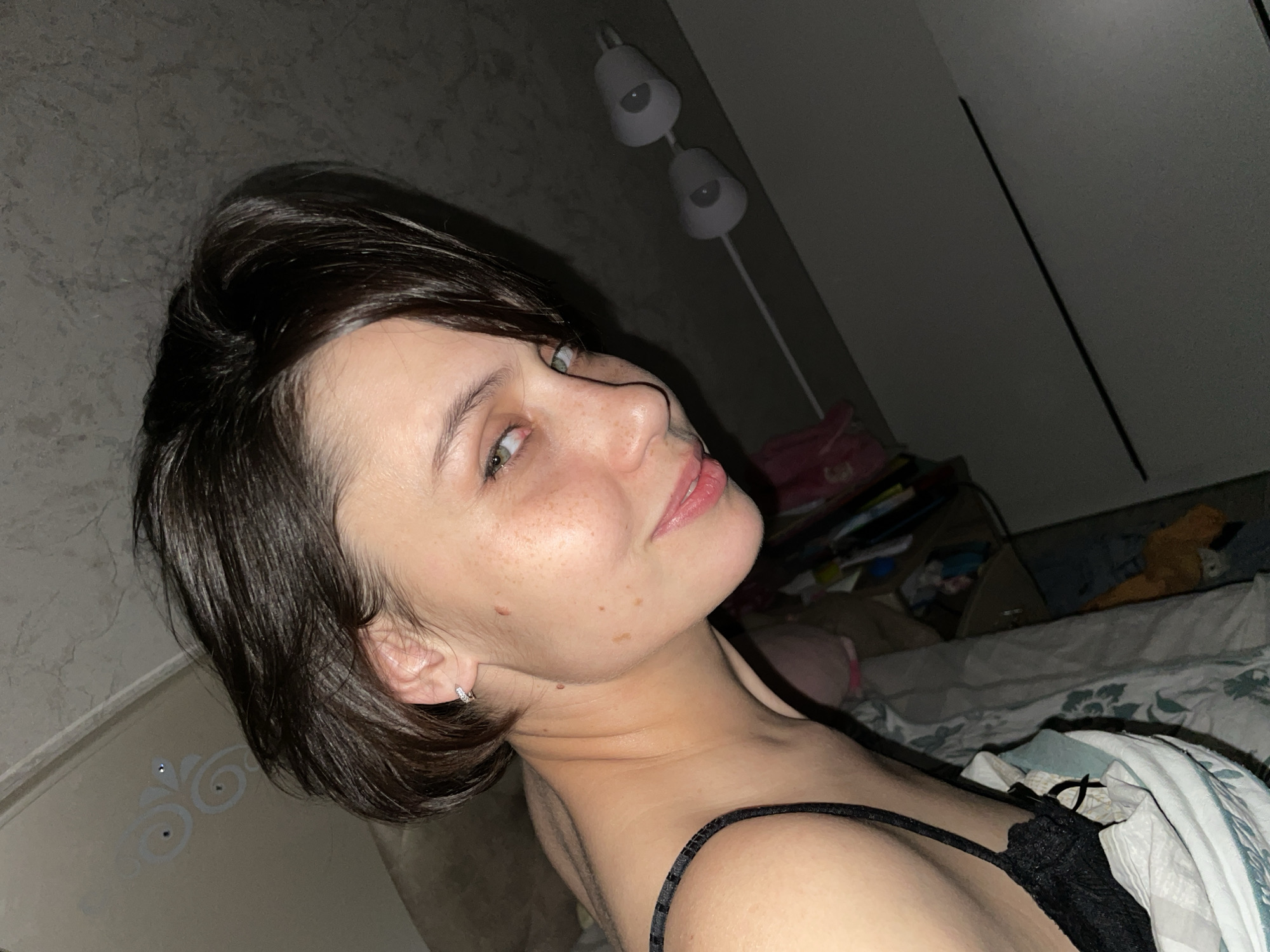 A woman with short brown hair, fair skin, and freckles is lying in bed. She is wearing a black tank top. The background includes a lamp with two lights, a closet or wall, and cluttered bedding. The location in the city of Izhevsk, Russia, cannot be determined from the image.