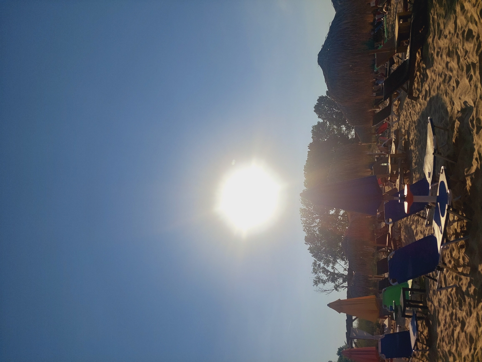 The image depicts a beach scene in Sozopol, Bulgaria. The sun is prominently featured in the upper portion of the sky, emitting a bright glare. In the foreground, beach chairs and umbrellas are arranged on the sand. Some chairs are occupied by people, though their faces are not clearly visible due to the lighting and distance. Trees and more thatched umbrellas line the background, suggesting a resort or populated beach area. The sand is golden and appears well-trodden. The overall atmosphere is one of a sunny day at the beach.