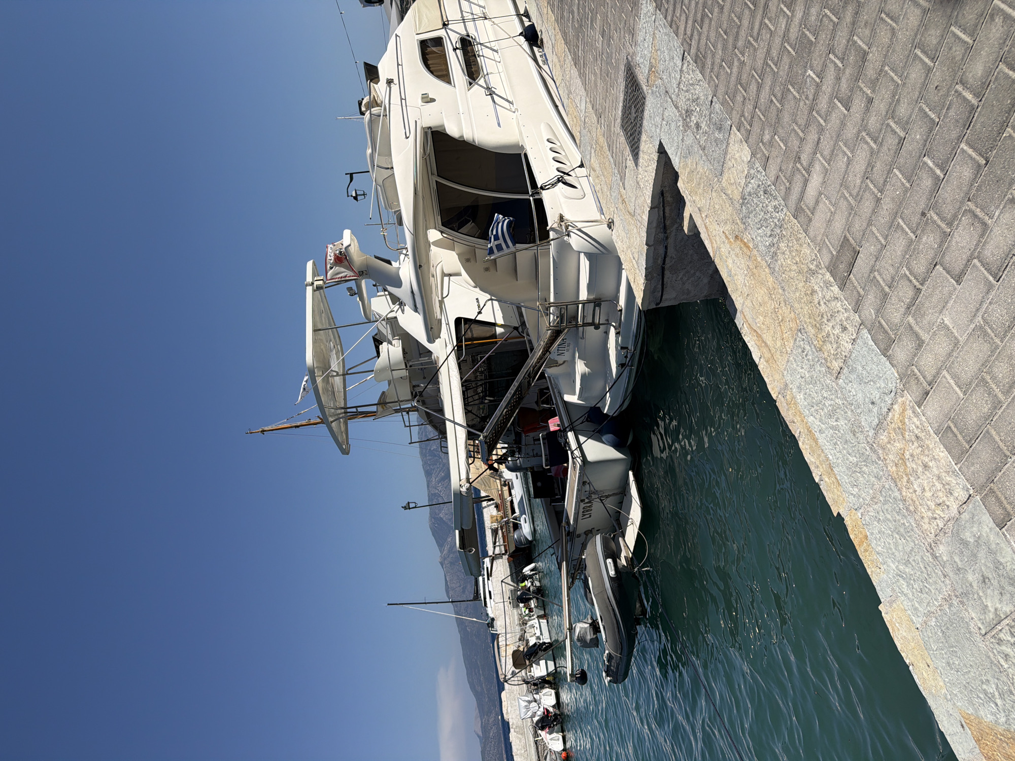 A wide-angle, low-angle shot captures several docked boats in a harbor under a clear blue sky. The focal point is a large white motor yacht, with a Greek flag prominently displayed on its stern. To the left, other smaller boats, including a rigid inflatable boat (RIB) dinghy, are moored. The boats are docked next to a stone quay with a paved walkway. In the background, mountains are visible against the horizon. The water is a deep blue-green color, with gentle ripples. The overall scene suggests a sunny day in a coastal area of Greece, likely in the Municipal Unit of Kalamos, given the presence of Greek flags and the mountainous coastal landscape. There are no people visible in the image.