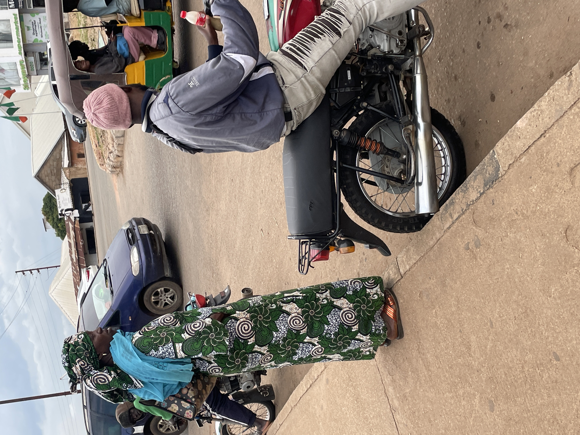 In Jos, Nigeria, a woman in a green and white patterned dress and headscarf, carrying a bag, stands on a sidewalk next to a parked motorcycle. A man wearing a pink beanie, grey jacket, and ripped light-colored pants rides another motorcycle in the foreground, holding a bottle of Coca-Cola. In the background, a blue minivan and a motorized rickshaw are visible on a street, with buildings and a supermarket in the distance. The scene is captured on an overcast day.