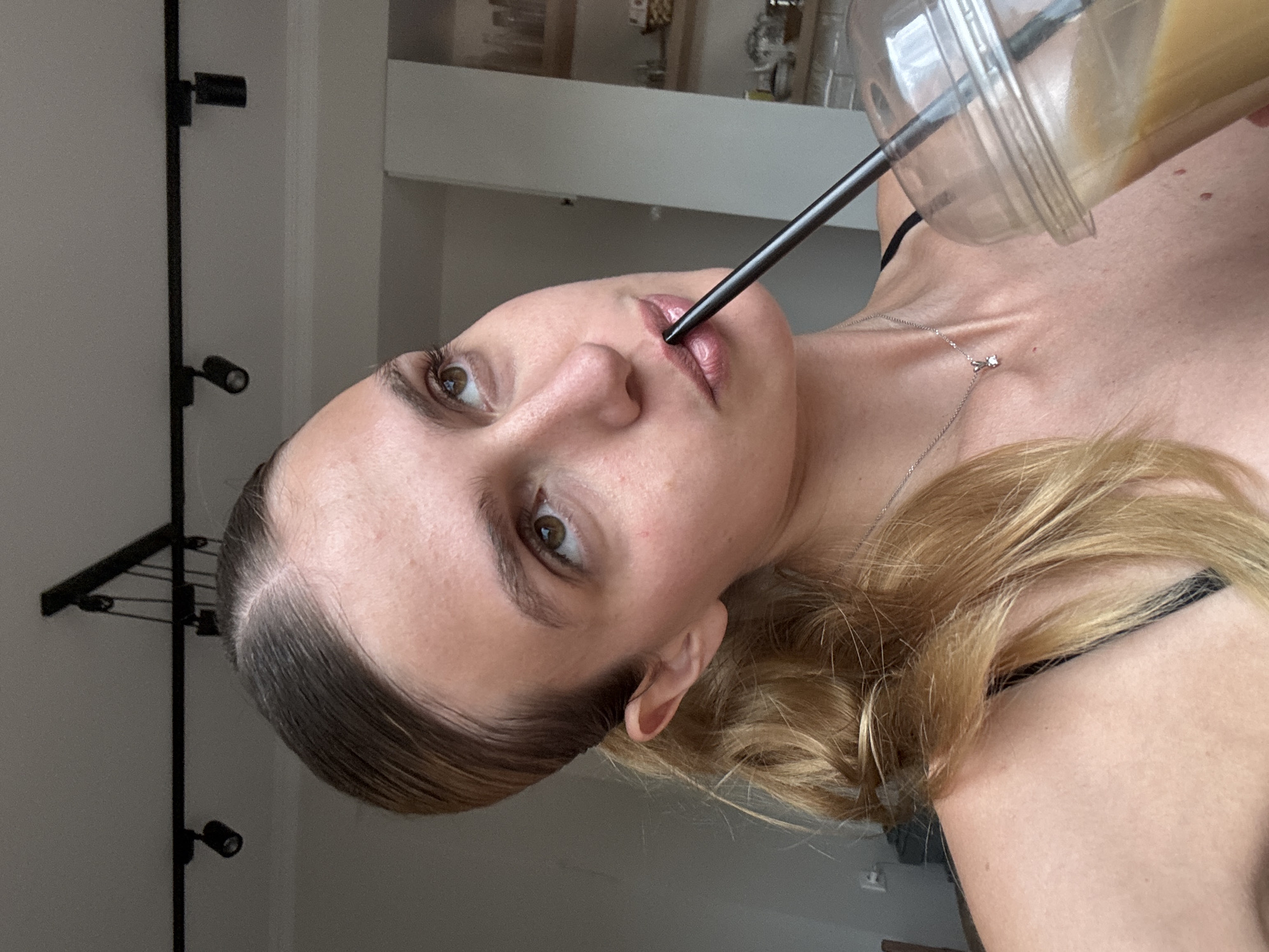 A woman is pictured drinking from a clear plastic cup with a black straw. She is fair-skinned with blonde hair pulled back and a silver necklace. The background includes a light fixture and a partially visible room with a white wall. The city and specific location are unknown.