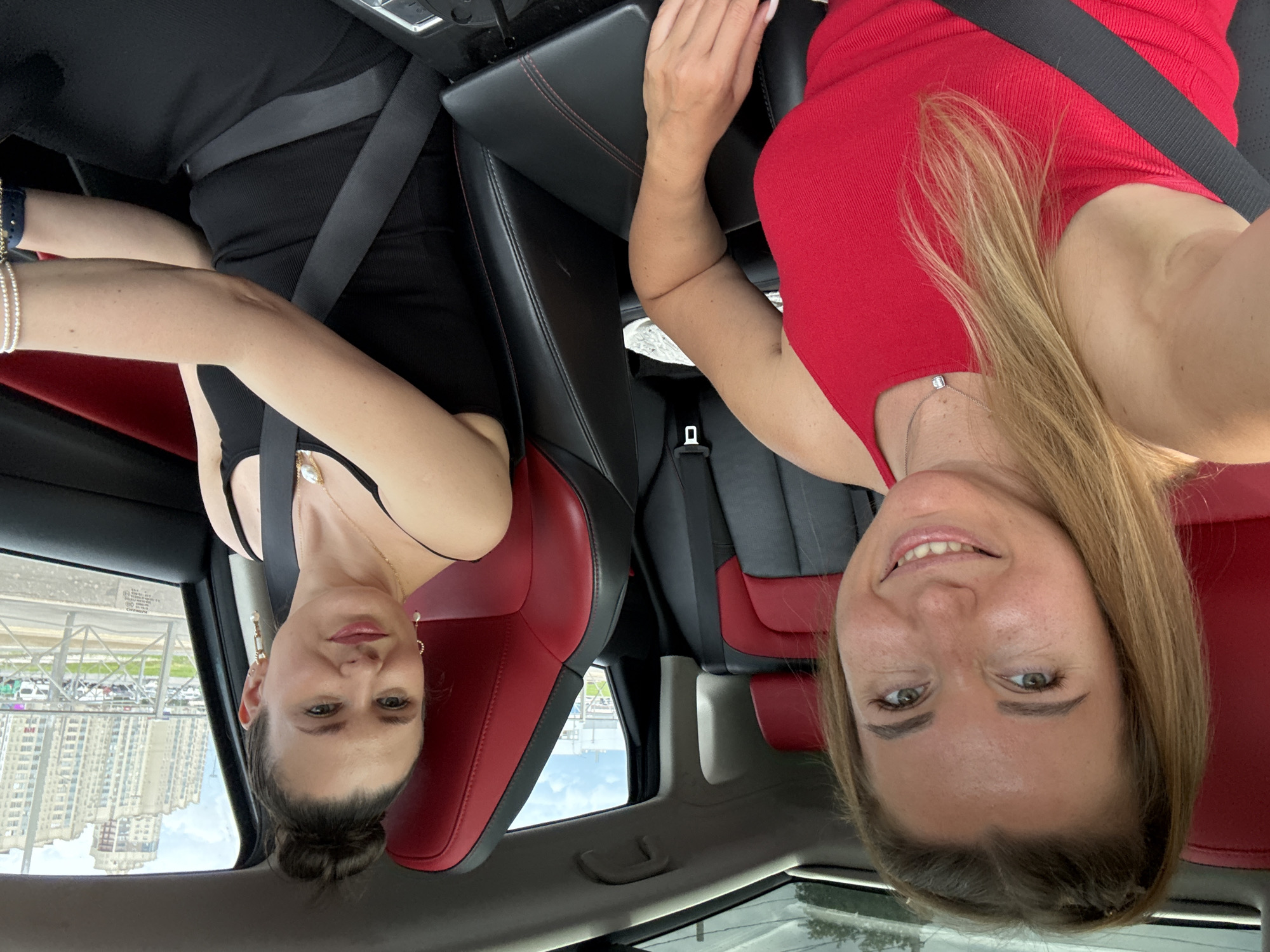 Two women are inside a car, with the camera positioned to capture both of them. The woman on the left, who appears to be taking the selfie, has blonde hair pulled back into a ponytail and is wearing a red sleeveless top. She is smiling at the camera. The woman on the right has her hair in a bun and is wearing a black tank top. She is looking towards the right side of the frame and has a slight smile. Both women are wearing seatbelts. The interior of the car features red and black seats. Outside the car window, a city skyline with tall buildings is visible, along with a road and some parked cars, suggesting an urban environment.