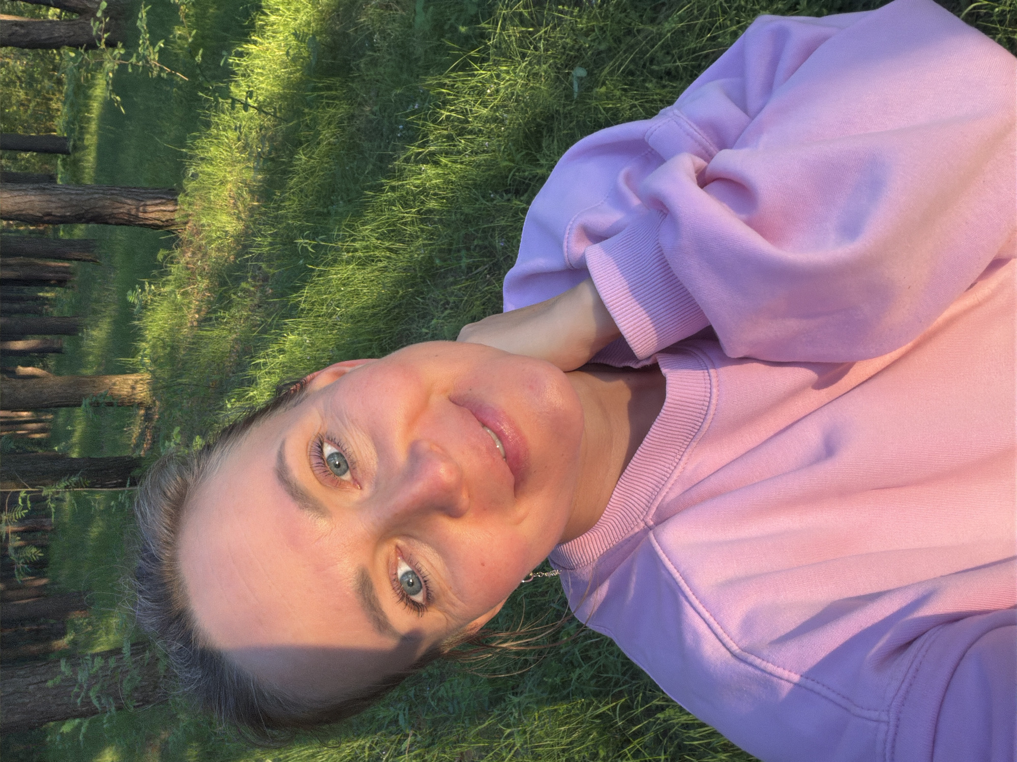 A woman takes a selfie while lying on her back in a grassy outdoor setting in Chaikovsky, Russia. She is positioned with her head towards the top of the frame, looking up at the camera with a gentle smile. Her blue eyes are open, and her face is illuminated by warm, golden-hour sunlight, suggesting either late afternoon or early evening. She is wearing a light pink long-sleeved sweatshirt. Her dark hair is visible around her head, blending with the natural surroundings.
The background consists of lush green grass, with several tree trunks rising vertically, indicating a park or wooded area. The sunlight casts long, soft shadows across the grass and partially on the woman's face. The overall mood is serene and relaxed, portraying a moment of leisure in nature. No text is visible in the image.