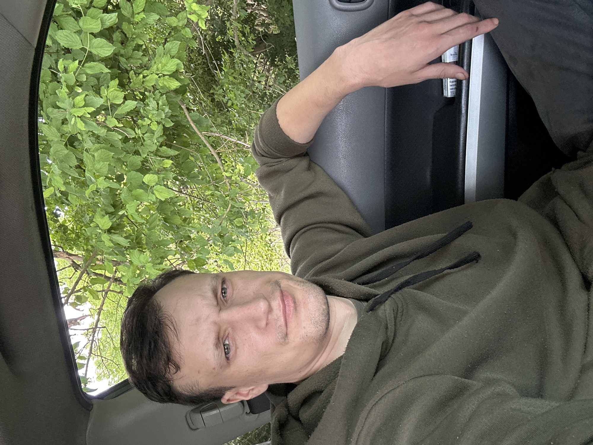 A man is seen from the chest up, sitting in the driver's seat of a car. He is wearing a dark green hooded sweatshirt. His left arm is resting on the car door. Outside the car window, green foliage is visible. The man's right hand is visible on the door panel and appears to be holding a small white and blue package. The interior of the car is dark gray. The scene is likely outdoors, possibly in a rural area given the presence of trees. No specific details about Makiv, Ukraine, are evident in the image.