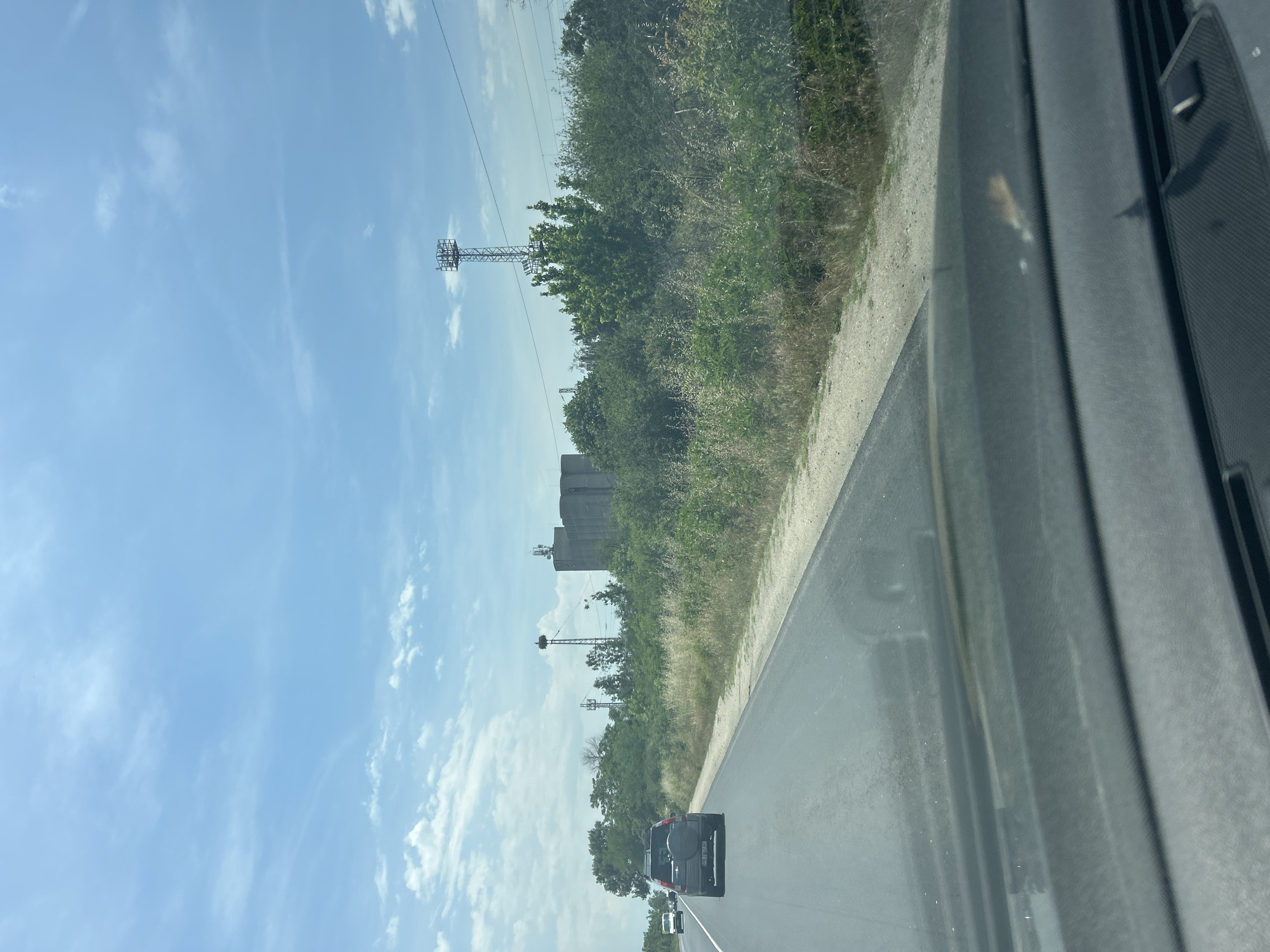 The image captures a bright, sunny daytime scene from the perspective of a moving vehicle, likely traveling through Dalgo Pole, Bulgaria. The view is primarily focused on the landscape alongside a road.
In the upper portion of the frame, a clear light blue sky is visible, adorned with scattered white clouds. Several tall, metal lattice structures, resembling communication or transmission towers, rise prominently above the treeline, standing against the sky. Below these towers and extending across the middle ground is a dense expanse of green trees and bushes. Partially obscured by the vegetation but clearly visible in the mid-ground are large, grey, blocky industrial buildings, which appear to be silos or a factory complex.
To the right and bottom of the frame, a two-lane asphalt road extends into the distance. A dark-colored SUV, possibly with a spare tire mounted on the rear, is seen traveling ahead in the left lane, with another lighter-colored vehicle further in the distance. A dirt or gravel shoulder runs alongside the road, separating it from the dense vegetation. On the far right and bottom edge, parts of the vehicle's dark-colored dashboard and windshield frame are visible, suggesting the photo was taken from inside a car. There are some reflections on the windshield.
The overall impression is one of travel on a highway or rural road during clear and pleasant weather. No discernible text is visible anywhere in the image.