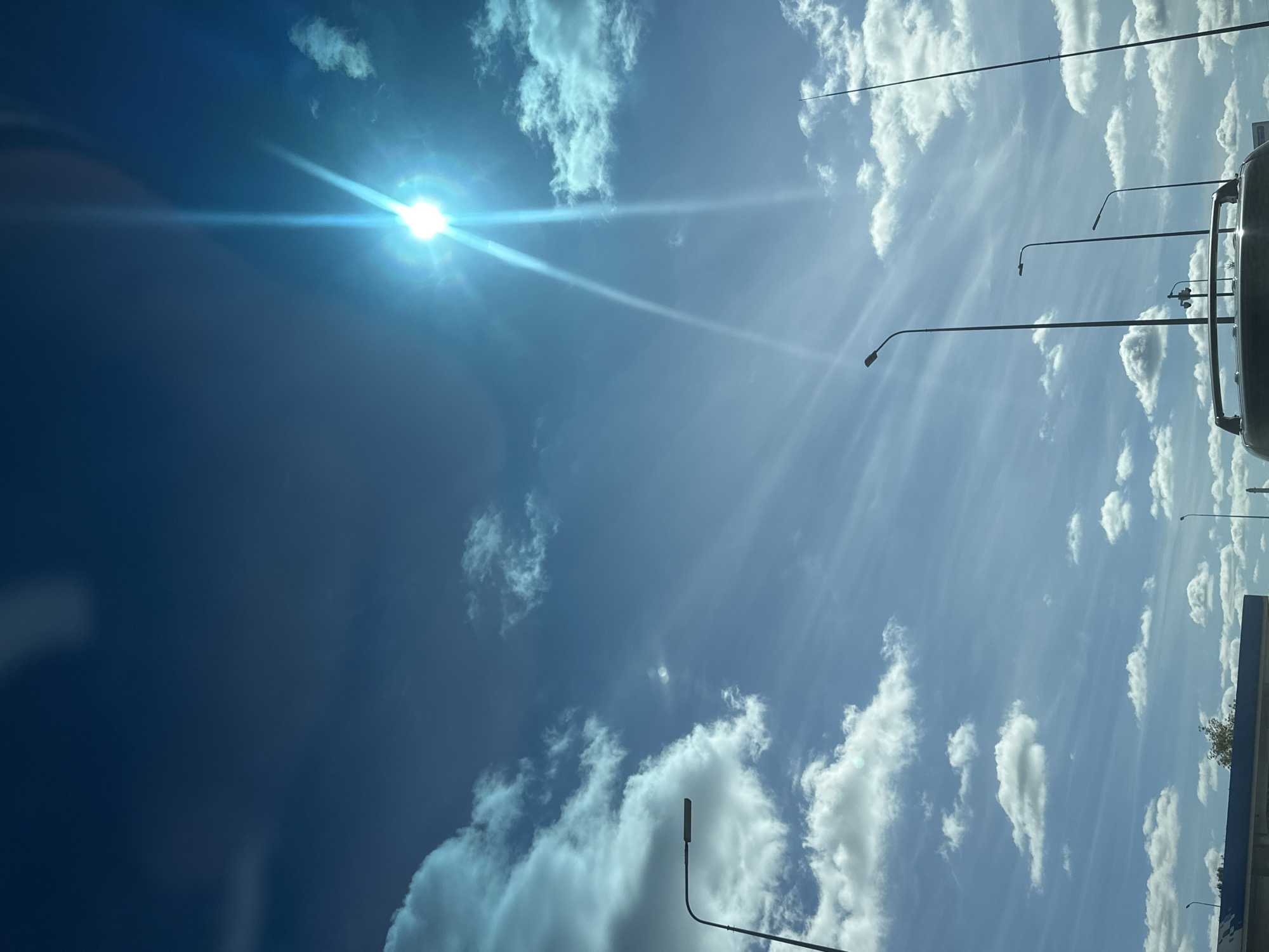 This is a vertical, medium-shot, eye-level photograph of a bright, sunny sky with scattered clouds and several street lamps. The sun is prominent in the upper right quadrant, emitting a strong lens flare that creates bright streaks of light. The sky is a deep blue, with wispy cirrus clouds interspersed with larger cumulus clouds, some of which appear tinged with blue from the sunlight.
In the foreground, the roof of a vehicle is visible at the bottom of the frame. Above the roof, a line of street lamps can be seen, varying in height and design, with the tallest on the far right. The street lamps are silhouetted against the sky. In the lower left corner, a building with a blue roof is partially visible, and a small tree with yellowing leaves is to the left of the building. The environment appears to be an urban or suburban setting, possibly a roadside.
The scene is static and focuses on the sky and its elements. There are no people or animals clearly visible. The lighting is bright and sunny, creating a strong contrast between the illuminated sky and the darker silhouettes of the objects. The overall impression is one of a clear, bright day. It is not possible to determine the specific location in Podolsk, Russia, from the visual information provided.