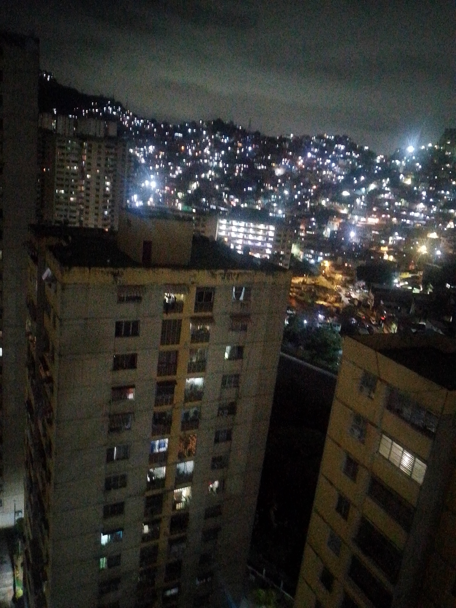 A nighttime aerial view showcases a densely populated hillside in Caracas, Venezuela, illuminated by countless lights from residential buildings and homes. The foreground features the side of a multi-story apartment building with visible windows, some lit from within. To the right, another apartment building, also partially lit, extends into the frame. The background is dominated by the sprawling urban landscape climbing the hills, with lights creating a twinkling effect against the dark sky. The scene depicts a vibrant, yet dimly lit, urban environment at night.