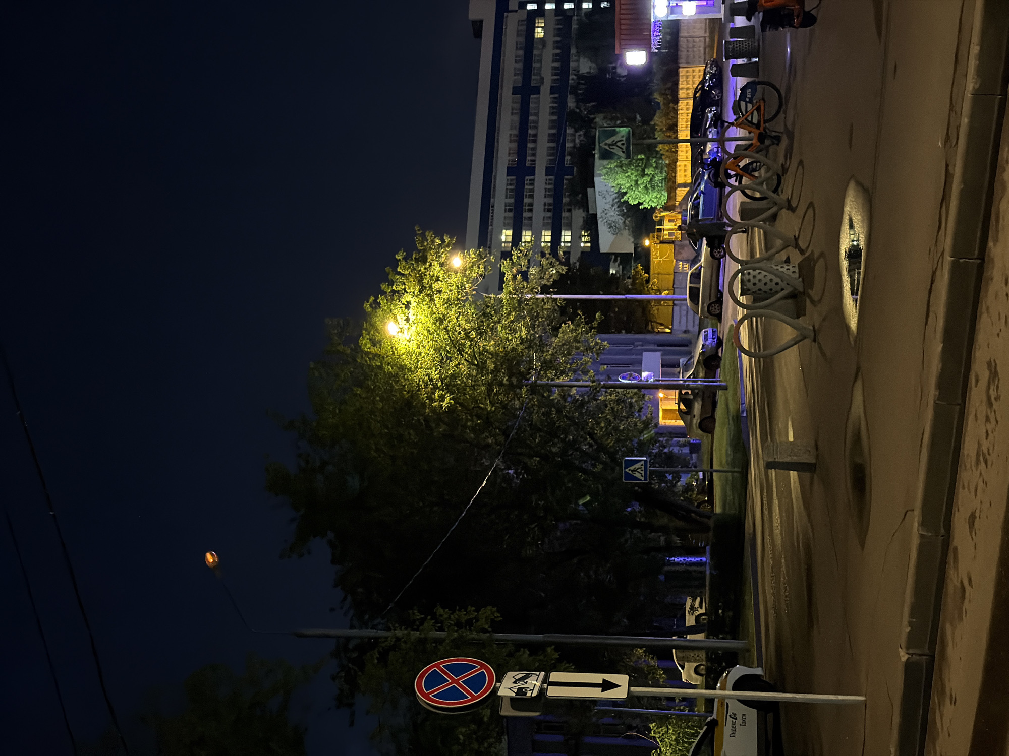 The image depicts a nighttime street scene in the city of Khimki, Russia. A streetlamp illuminates a tree and a portion of the road. In the foreground, a concrete curb separates the sidewalk from the street. The street features a marked crosswalk and a bicycle rack with several bicycles. Cars are parked along the side of the road. In the background, a multi-story building with illuminated windows is visible. A taxi with the inscription "Яндекс Такси" (Yandex Taxi) is parked on the left side of the image. Traffic signs, including a no-parking sign and a pedestrian crossing sign, are present. The sky is dark, indicating it is night.