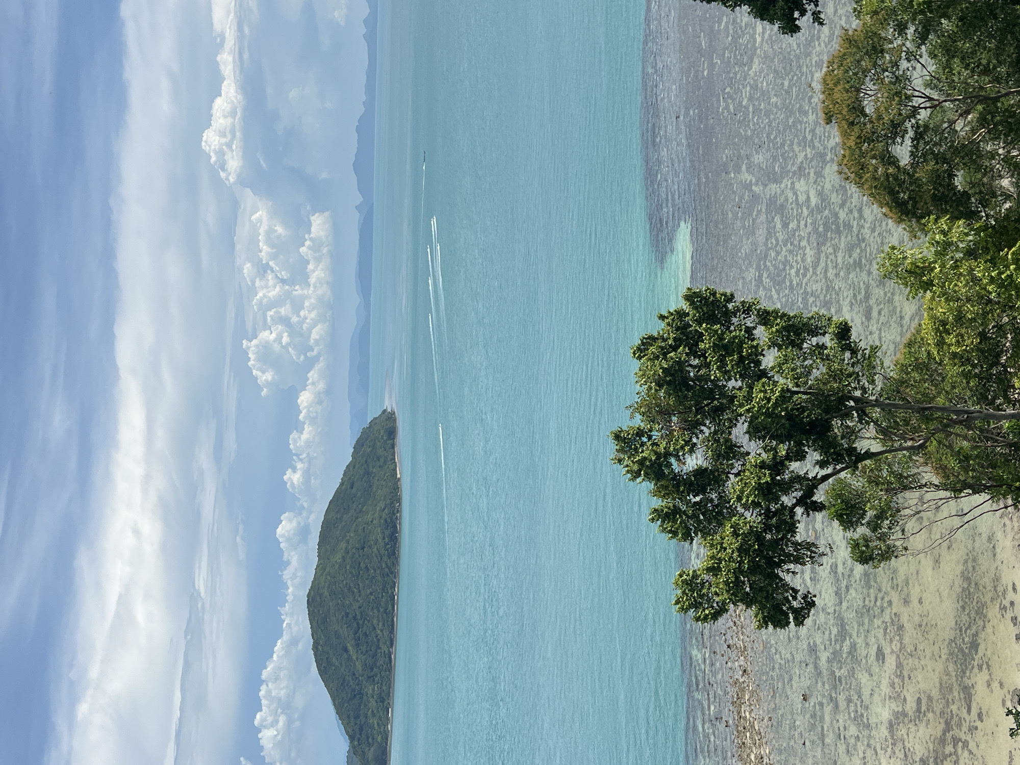 The image captures a scenic view of the ocean and a lush green island from a high vantage point. The foreground features the tops of several trees with dense green foliage, framing the view of the water. The ocean itself is a vibrant turquoise, with areas of shallower water closer to the shore revealing a rocky or coral seabed. Several speedboats are visible on the water, leaving white wakes as they travel. In the distance, a large, rounded island covered in dense vegetation sits on the horizon, with a scattering of smaller islands or landmasses beyond it. The sky is filled with a mix of blue sky and large, fluffy white clouds, suggesting a partly cloudy day. The overall impression is of a tropical coastal landscape. The location is identified as Baan Thong Krut, Thailand.