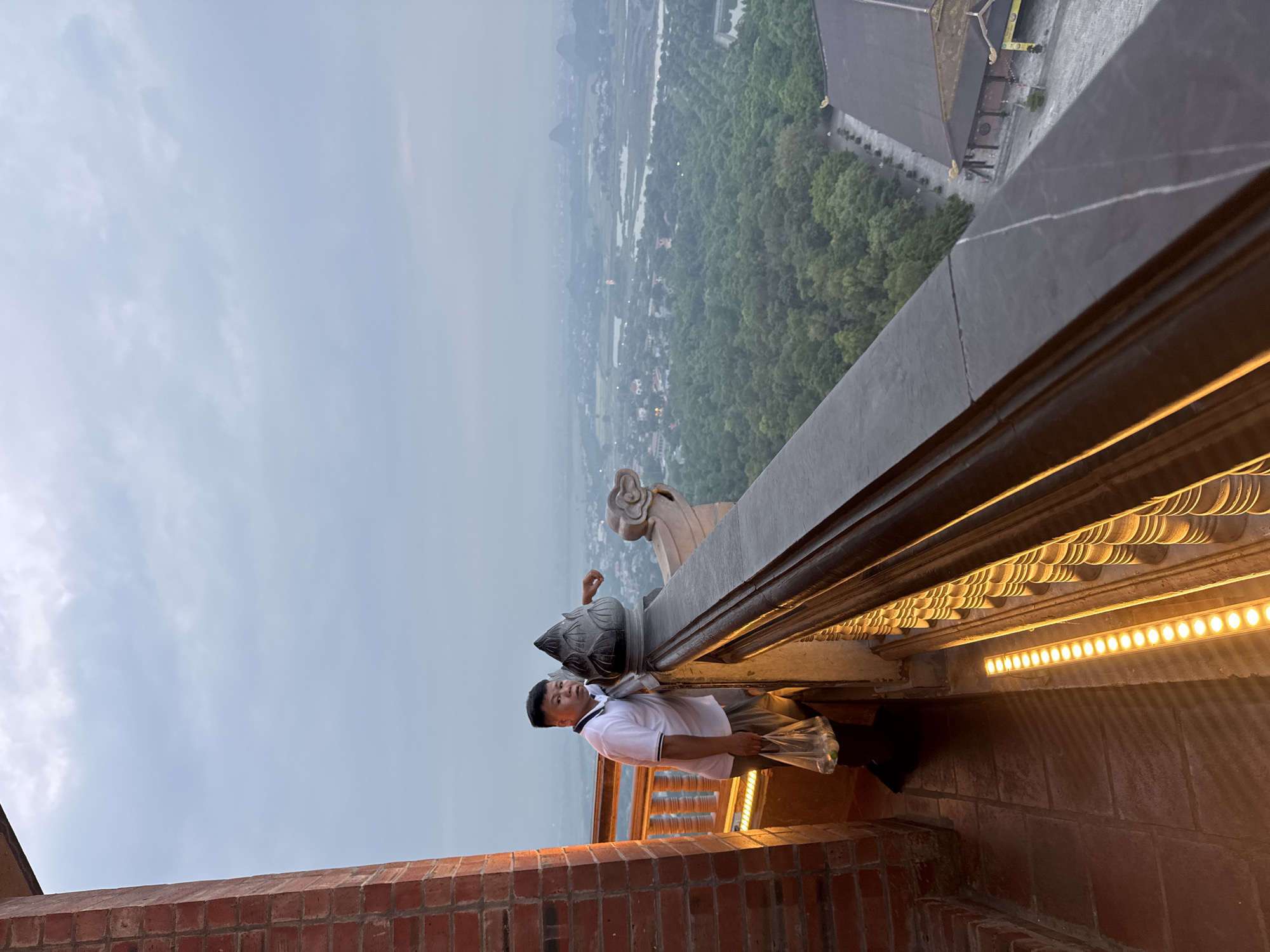 A man stands on a balcony overlooking a valley at dusk in Tây Hoa Lư Ward, Vietnam. The man is wearing a white polo shirt and brown pants, and he is holding a plastic bag. He is leaning on the balcony railing, which is made of dark stone and has decorative wooden balusters. The balcony is lit by a row of yellow lights. In the distance, the valley is covered in trees, with a few houses and bodies of water visible. A temple with a distinctive roof can be seen in the lower right corner of the frame. The sky is overcast and the light is dim.