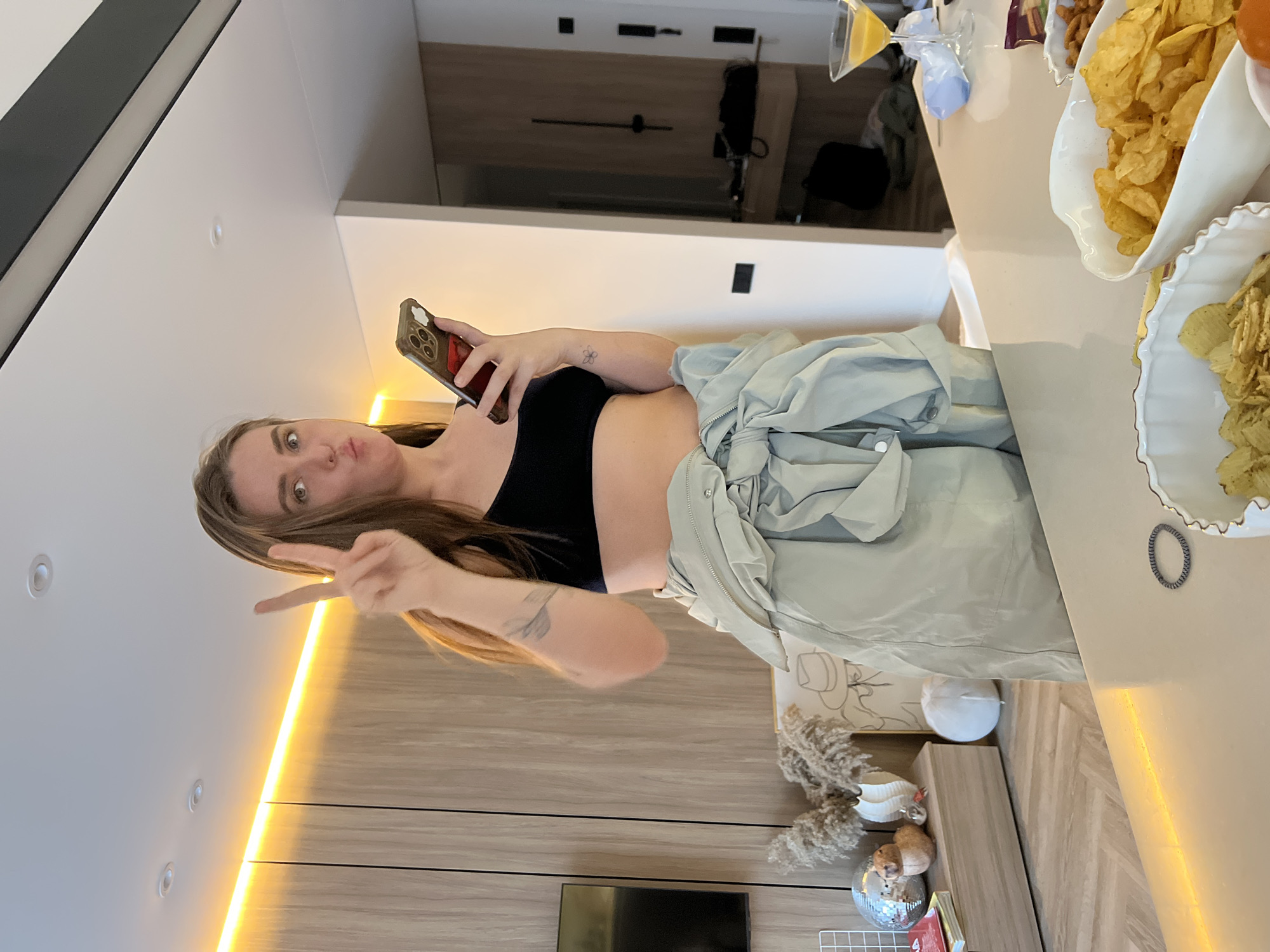 A woman in a black top and light green pants stands in a room, holding a phone up in her right hand and making a peace sign with her left hand. The room has light wood paneling on the walls, a black television, and a light strip running vertically on the wall. Next to the woman is a table with bowls of chips and a drink. Рубцово, Russia is not visibly depicted in the image.