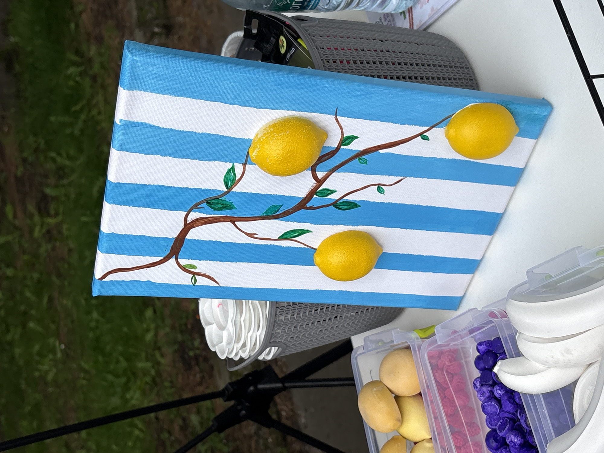 The image depicts a close-up view of a painting of lemons on a striped canvas, positioned on a white table outdoors. The painting features a brown branch with green leaves, from which three yellow, textured lemons hang. The canvas has vertical blue and white stripes.
To the left of the painting, several plastic containers hold various items: one contains yellow, lemon-like objects; another holds small, pink and purple objects; and a third holds white, banana-shaped objects. Behind the painting, a gray basket is filled with stacked white paper plates. A black tripod is visible in the background, partially obscured by the basket.
To the right of the painting, another gray basket holds a clear plastic bottle of water and some black packages. The background is a blur of green grass and foliage, suggesting an outdoor setting. The lighting indicates it is daytime. The scene is photographed from a slightly elevated angle.
There are no visible people in the image. The overall impression is of an art display or crafting activity, possibly at an outdoor market or event. This scene does not provide any specific clues to its location in Birobidzhan, Russia.
