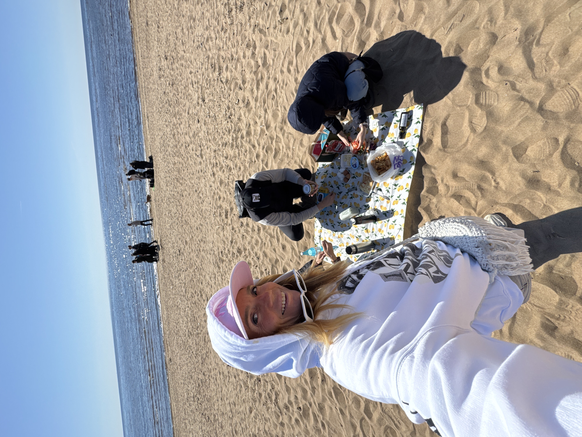 The image shows a sunny beach in Komarovo, Russia. A woman is taking a selfie, wearing a white hoodie and a pink hat. Behind her, two people are sitting on a picnic blanket with lemon print. The blanket has food items like tomatoes and chips. There are also bottles of water and a thermos. Further back, there are other people walking on the beach near the water. The sky is clear and blue. The sand is light-colored.
