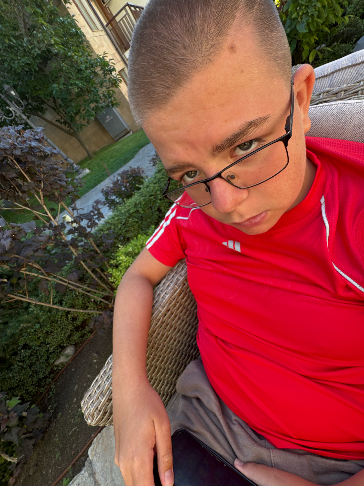 In Sveti Vlas, Bulgaria, a young person, possibly a boy, sits outdoors in a wicker chair, wearing a red t-shirt with white stripes and black-rimmed glasses. The individual is holding a dark-colored smartphone. The background features lush green foliage, a building with a balcony, and a paved pathway. The lighting suggests it is daytime, possibly late afternoon or early evening, with sunlight casting shadows.