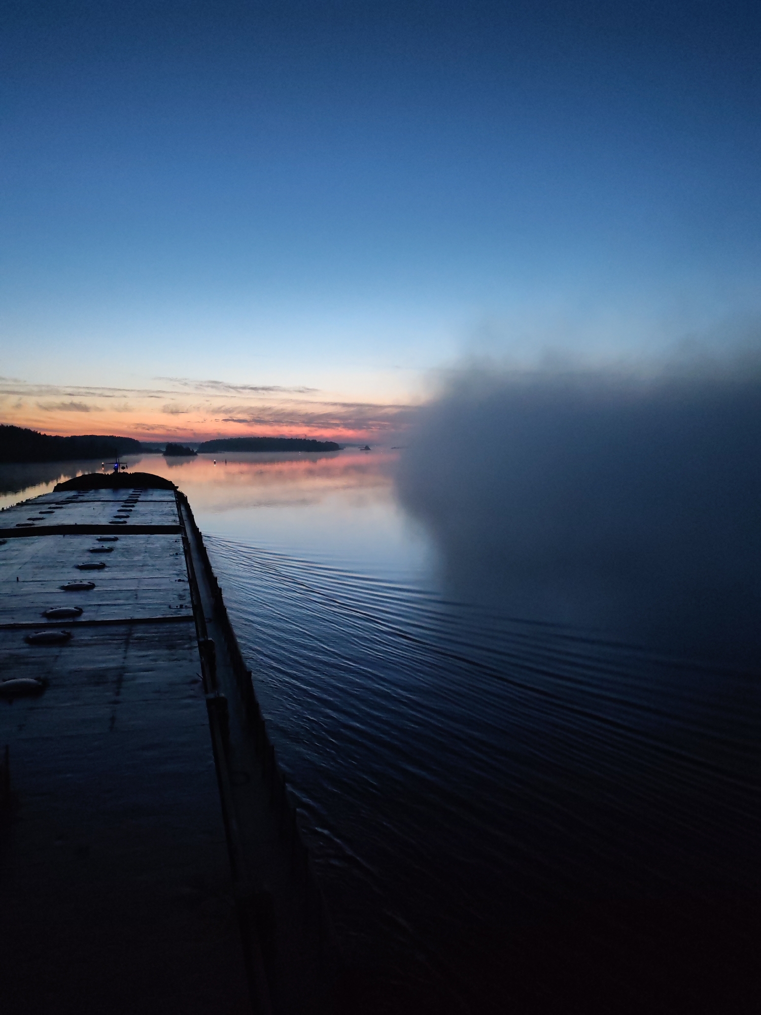 A tranquil, wide-angle shot taken from the deck of a barge or boat at dawn or dusk, possibly in Poteryaevo, Russia, but the location is not definitively confirmed. The sky transitions from a deep blue overhead to a soft orange-pink at the horizon. In the distance, silhouetted islands and landmasses rest on the water's surface. To the right, a dense fog bank obscures the view. The water is calm, reflecting the sky's colors, and marked by the vessel's wake. The deck of the vessel is dark and wet, leading the viewer's eye into the distance. There are no people visible.