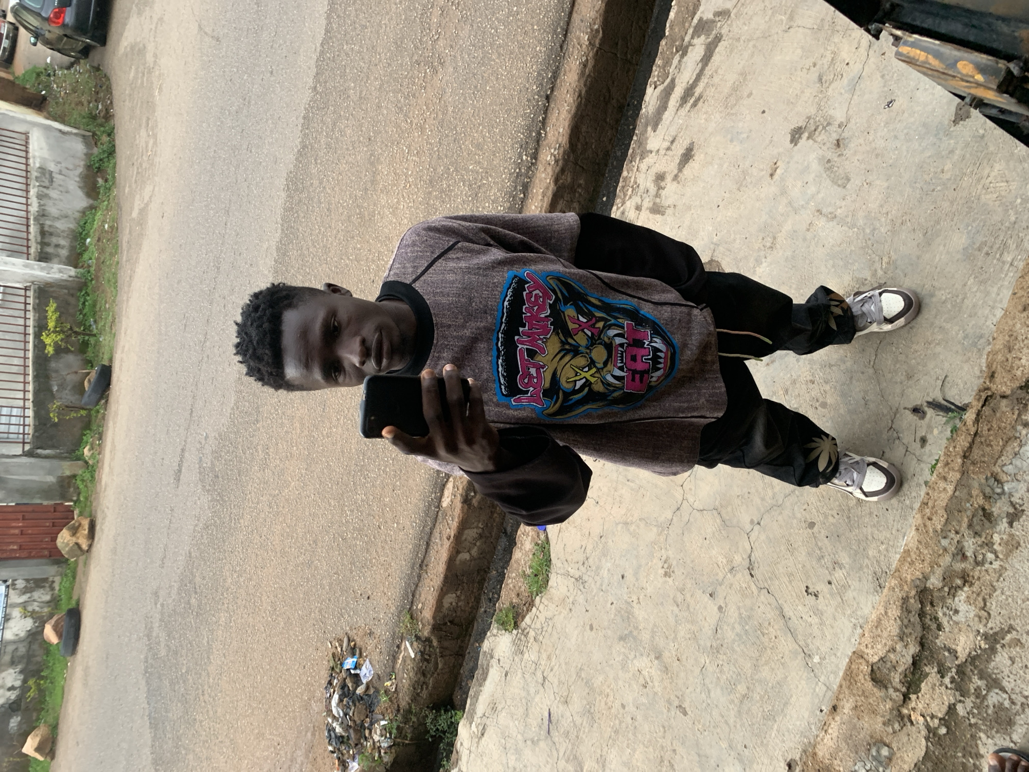 A young Black male is standing on a concrete sidewalk, holding a smartphone in his right hand and taking a selfie. He is wearing a gray long-sleeved shirt with a graphic print of a snarling animal face on the front, black pants, and white and gray sneakers. He has short, dark, curly hair. Behind him, a paved road is visible with a dirt shoulder and a drainage ditch. There are scattered pieces of debris on the shoulder, including plastic bags and what appears to be trash. Rocks and a deflated tire are also present on the side of the road. In the background, a building with a red metal door and a fence can be seen. The environment appears to be an urban setting in Jos, Nigeria.