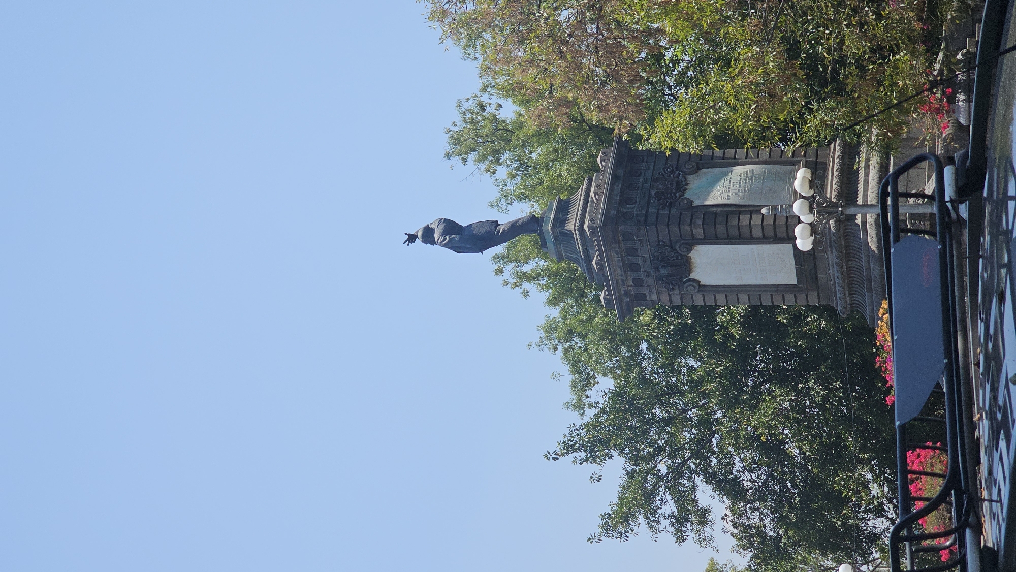 The image displays a grand public monument set against a clear, bright blue sky. A bronze statue of a distinguished man stands prominently atop a tall, ornate stone pedestal. The figure is depicted in formal attire, including a coat, with his right arm extended and hand pointing forward, suggesting a gesture of command or oration.
The multi-layered pedestal supporting the statue is intricately carved with architectural details and decorative elements. It features at least two rectangular plaques of a lighter color, affixed to its sides. While some faint inscription is visible on the upper plaque, the text is not decipherable.
Lush green trees with some scattered autumnal leaves frame the monument, hinting at a park or public square setting. To the right, a classic lamppost with multiple white globe lights illuminates the area. Dark metal railings, possibly part of a fence or barrier, are partially visible in the foreground, with vibrant red and yellow/orange flowering plants adding splashes of color near their base. The scene is illuminated by daylight, indicating clear and sunny weather. The monument reflects historical significance, typical of the cultural landscape of Puebla, Mexico.
