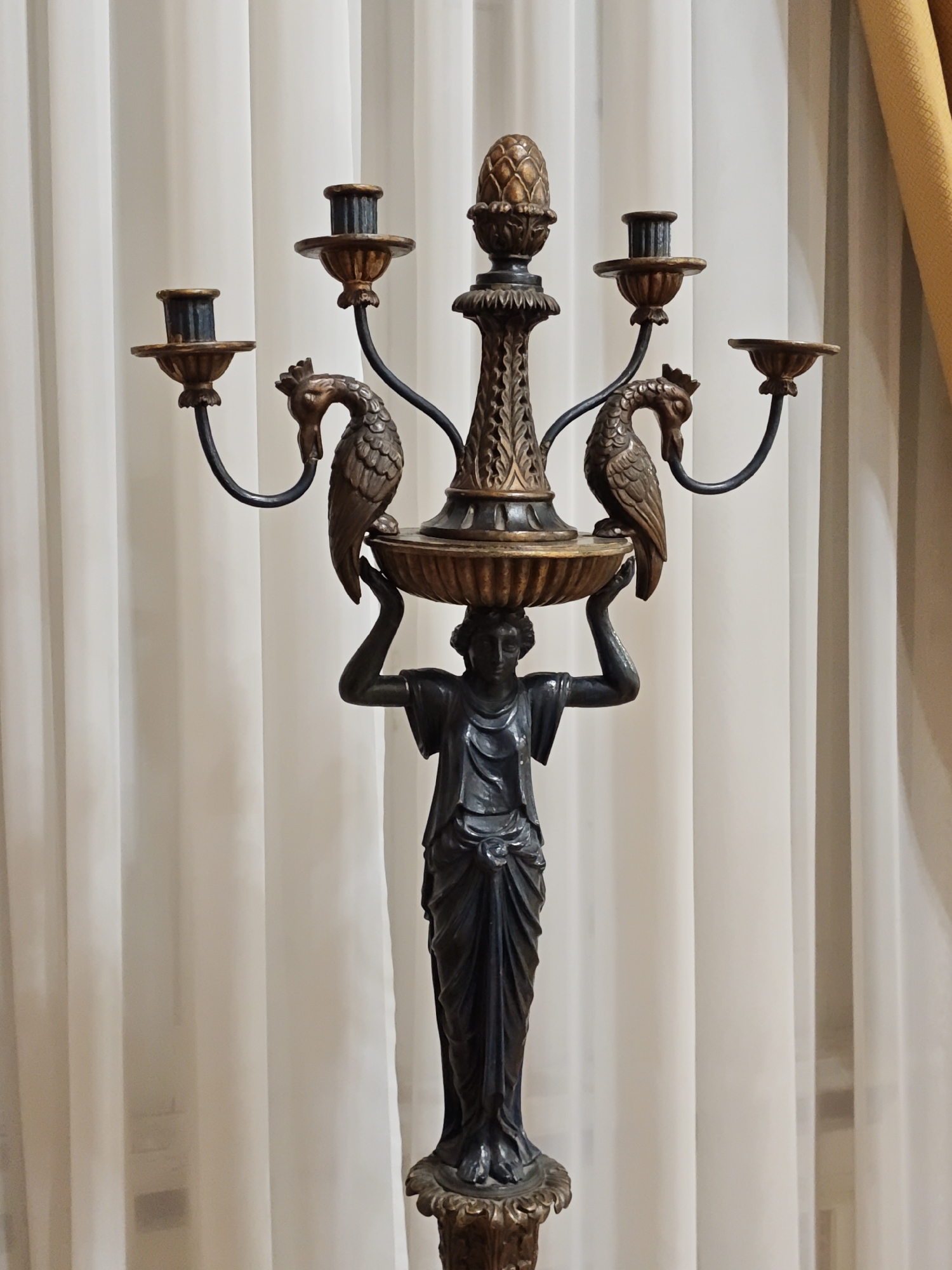 The image shows a close-up, eye-level, frontal view of an ornate, dark bronze candelabra standing in front of white sheer curtains. The candelabra is supported by a dark, stylized female figure with draped clothing, who holds a decorative bowl above her head. From this bowl, a central stem rises, adorned with carved leaf motifs and topped with a golden finial resembling a pinecone. Extending outwards from the stem are five arms, each curved and ending in a candle holder. Two of these arms are decorated with detailed, carved bird figures with distinctive crests, positioned to face outwards. The candle holders themselves are shallow cups with flared edges, some appearing to be dark blue or black inside, others showing a golden hue. The base of the candelabra is partially visible, appearing to be ornate and possibly gilded. The lighting suggests a soft indoor environment, casting subtle shadows. The background is dominated by the texture of the vertical folds of the white curtains, with a hint of a yellow-gold curtain visible on the right side. There are no people visible in the scene other than the sculpted figure. The location of this candelabra in Lviv, Ukraine, is not discernible from the image itself.