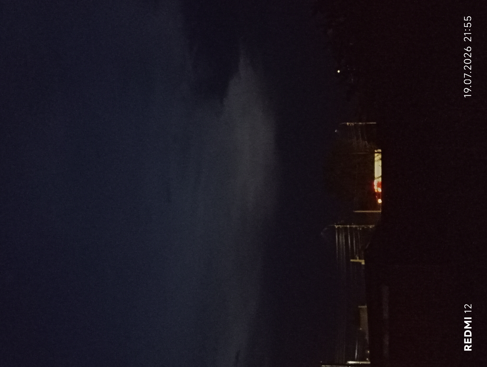 The image captures a dark, cloudy sky over what appears to be a semi-urban or suburban area at dusk or night. The lighting suggests it is nighttime, with artificial lights visible in the distance. In the foreground, a dark silhouette of land or structures is present. There are vertical elements that could be utility poles or streetlights. In the middle ground, a horizontal light source, possibly from a vehicle or building, is illuminated. The bottom left of the image shows the text "REDMI 12", and the bottom right displays the date "19.07.2026" and time "21:55". The location is identified as Smila, Ukraine. No people or specific actions are discernible due to the low light and distance. The environment is characterized by darkness and atmospheric conditions indicated by the clouds.