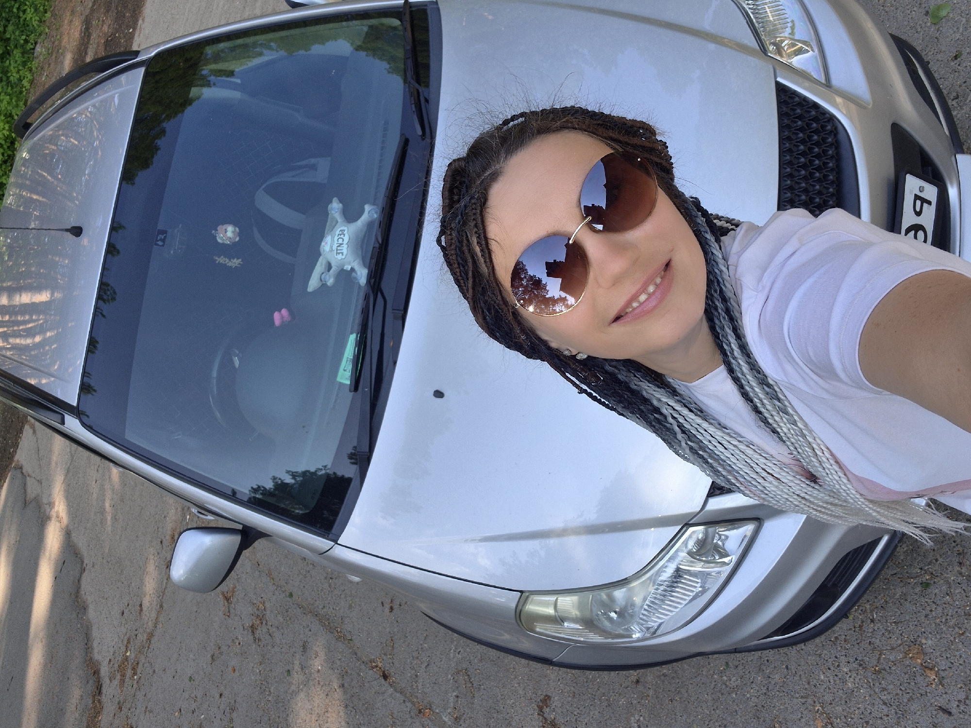 A woman with braided hair and sunglasses is taking a selfie in front of a silver car. The car is parked on a paved surface, likely a road or parking lot, with trees and foliage visible in the background, casting shadows. A license plate is visible on the front of the car, with Cyrillic characters that appear to be from Russia. The overall scene suggests an outdoor setting in a temperate climate. The location is identified as Chaikovsky, Russia based on the license plate.