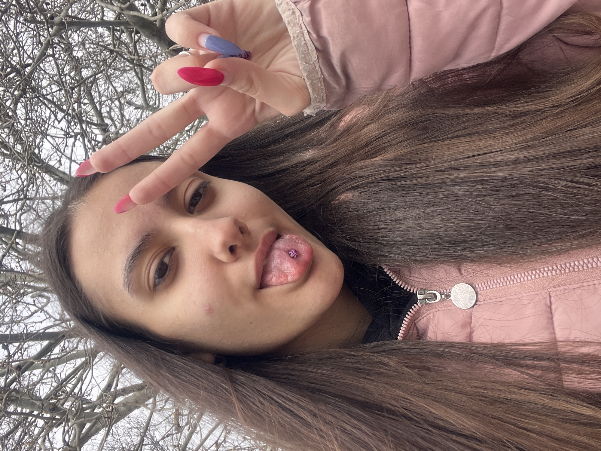 An upward-angled, close-up photograph depicts a young female outdoors in Varna, Bulgaria. The subject has long, dark brown hair, partially obscuring a pink puffer jacket and a black garment underneath. She is seen sticking out her tongue, which displays a purple, flower-shaped piercing. A small reddish blemish is visible on her right cheek below her eye. Her left hand is raised, forming a "peace" sign with the index and middle fingers. The fingernails are long and shaped, with the index and middle fingers painted bright pink, and the ring and pinky fingers featuring a blue-to-clear ombré with purple accents. The pink puffer jacket has a visible zipper with a circular, silver-toned charm attached. The background consists of numerous bare tree branches against an overcast sky, indicating an outdoor setting during a leafless season.