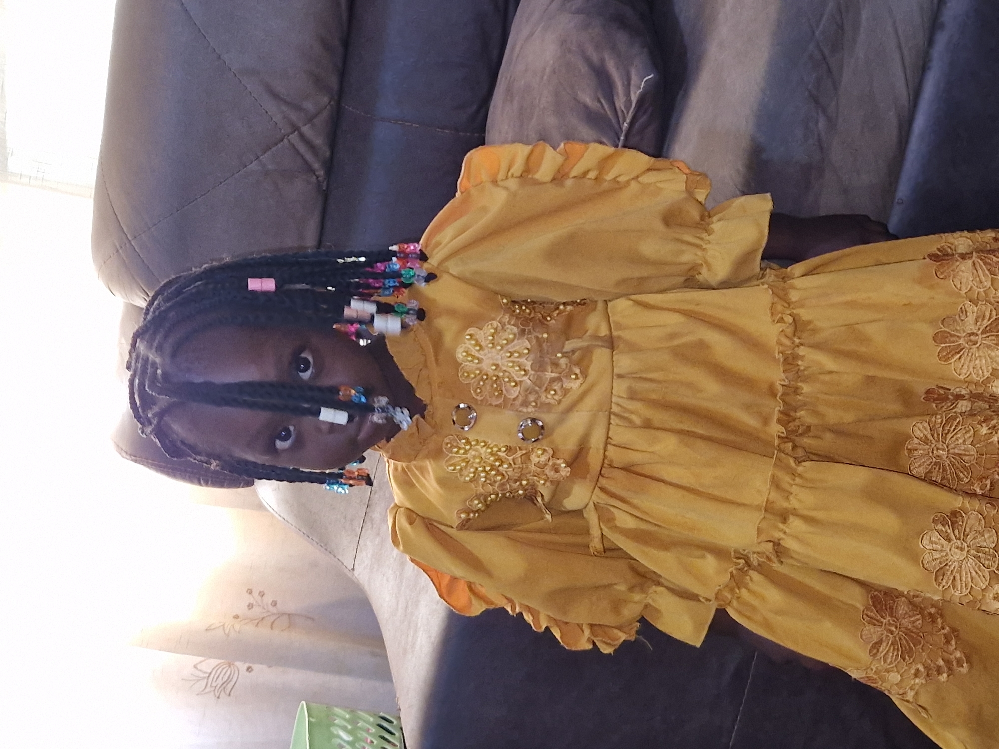 A young girl with dark skin, wearing a golden dress adorned with beads and lace, stands in front of a dark brown couch. Her braided hair is decorated with colorful beads. The background shows a sheer curtain with a floral pattern and a green basket to the left. The scene appears to be indoors, possibly a living room in Jos, Nigeria.