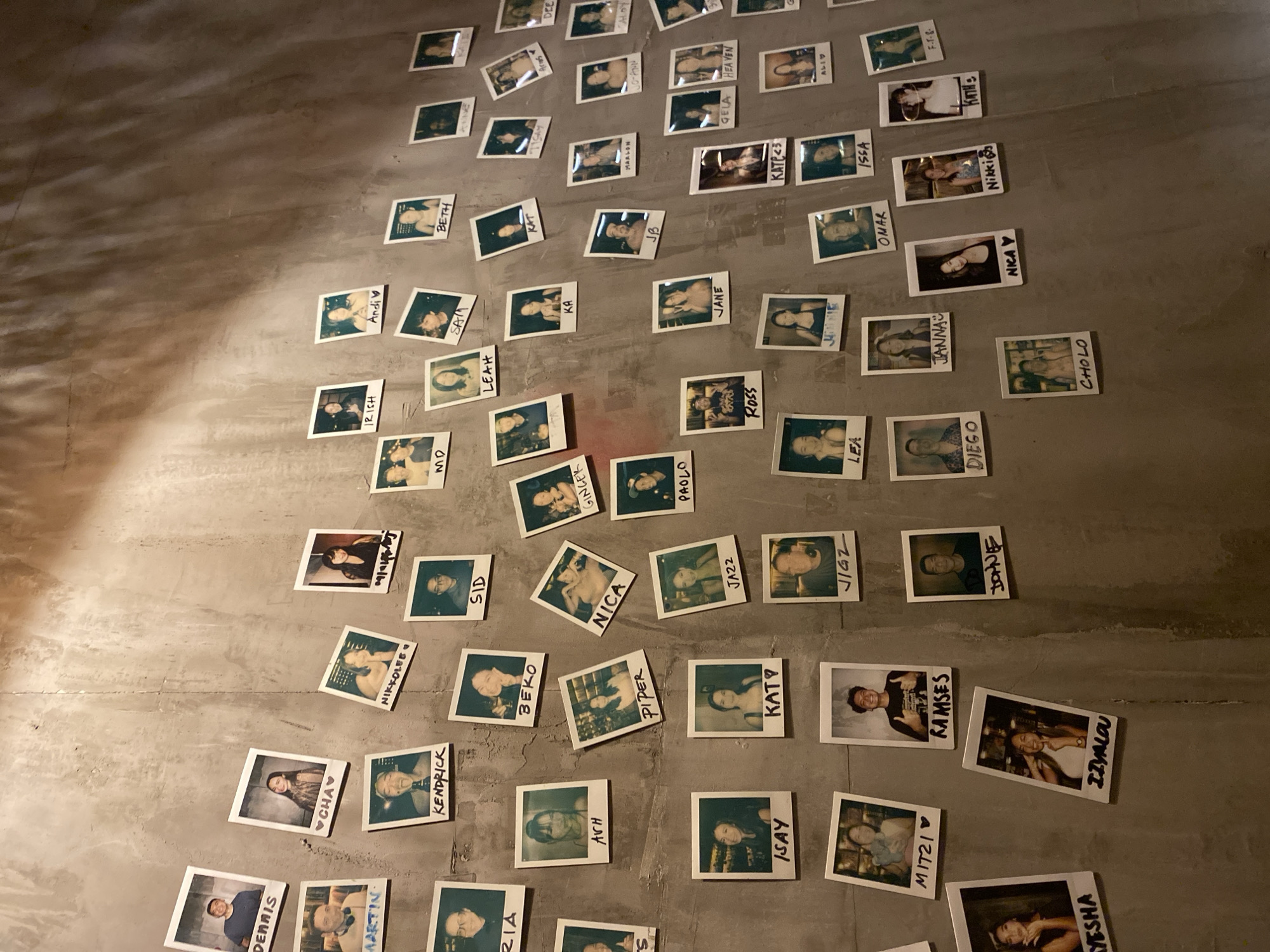 The image depicts a textured concrete wall adorned with numerous small, polaroid-style photographs. Each photograph features an individual, and most are labeled with handwritten names on white borders. The photographs are arranged in a somewhat haphazard grid pattern, with some overlapping slightly. The lighting suggests an indoor environment, possibly a bar or a private space, with shadows cast across the wall by the overhead lighting. The presence of a handwritten sign saying "DAVAO CITY" in the original image, along with the specific style of photographs and the overall ambiance, indicates a personal display within Davao City, Philippines. The subjects in the photographs appear to be diverse in age and gender, suggesting a collection of friends, family, or acquaintances. The wall itself is a neutral gray, providing a stark backdrop for the colorful and varied images.