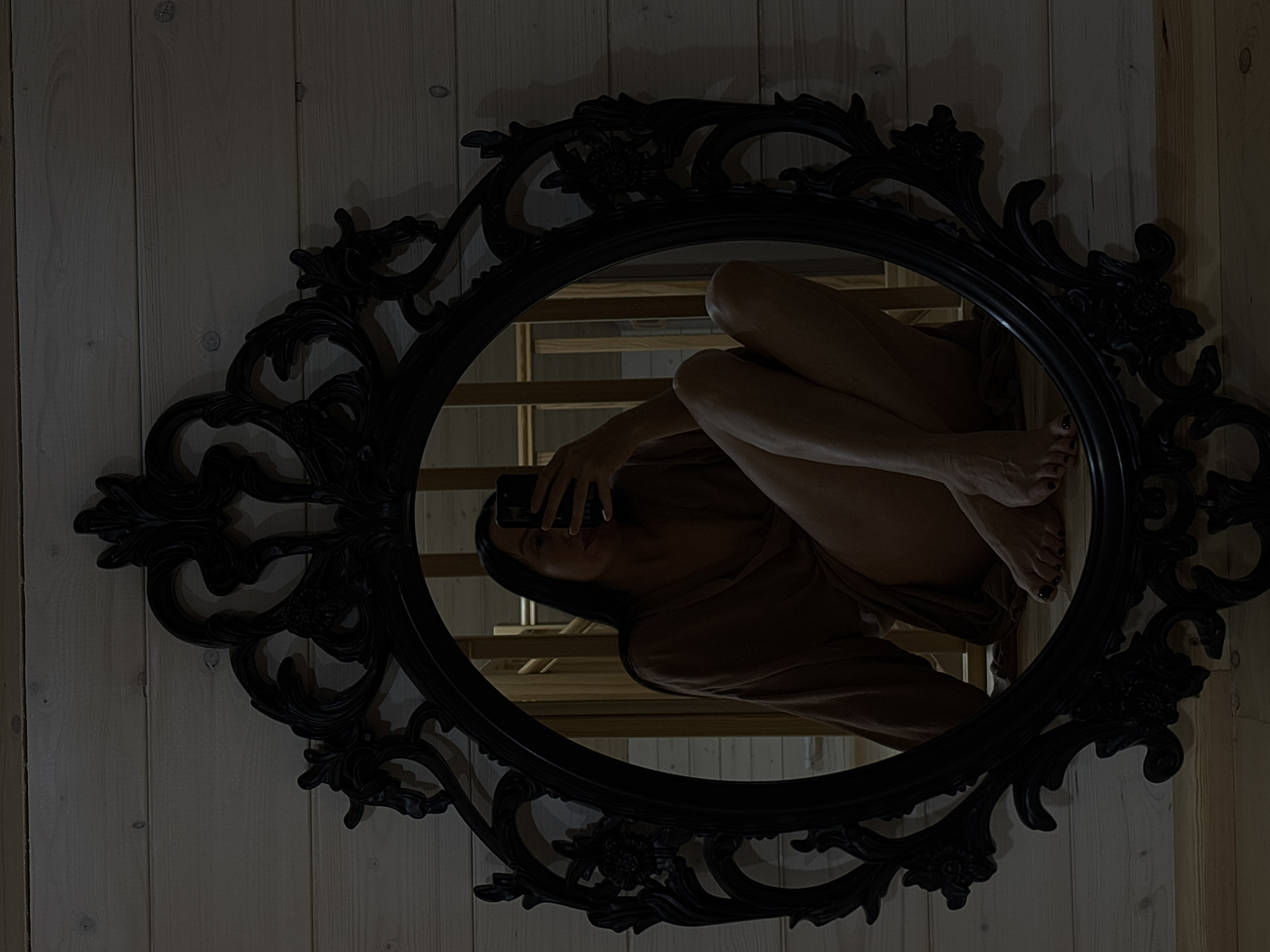 The image shows a woman reflected in an ornate, oval-shaped black mirror. The mirror is hanging on a wall composed of vertical wooden planks. The woman is holding a smartphone, presumably taking a photo of herself in the mirror. Her legs are bent, and one foot is visible. Behind her reflection in the mirror, a set of wooden stairs can be seen. The scene is dimly lit. It is impossible to determine if this image was taken in the city of Chaikovsky, Russia.
