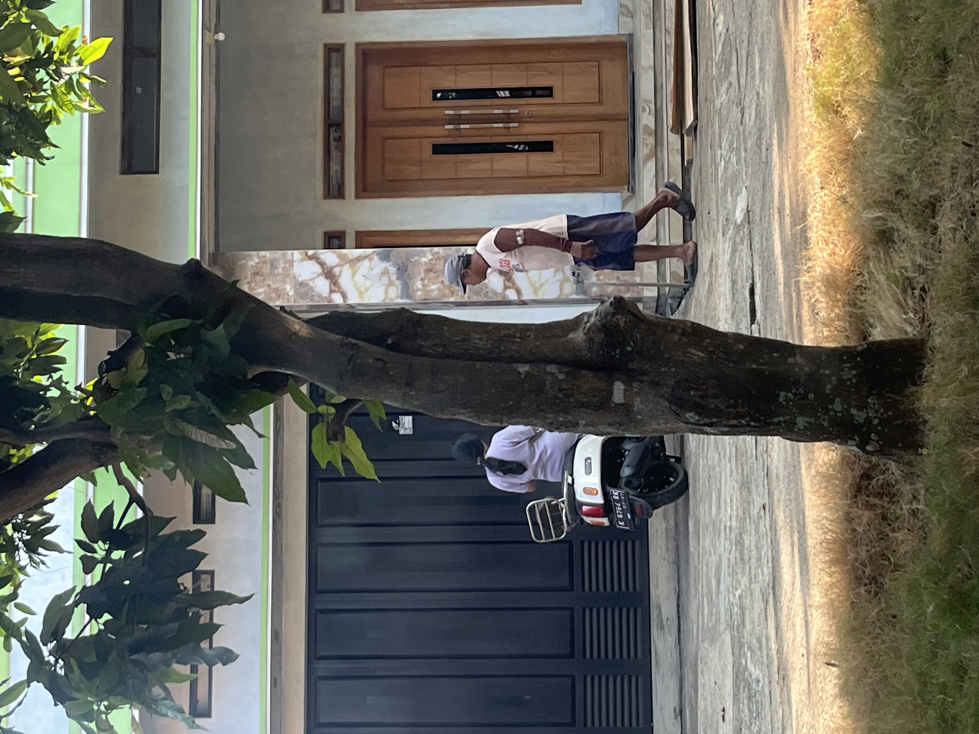 In Glagah Kulon, Indonesia, a man wearing a cap, light brown shirt with dark sleeves, and dark blue shorts walks on a paved surface. He has a bandage on his upper left arm. To his left, a white scooter is parked, and a person wearing a light purple shirt and dark pants is seated on it, facing away from the camera. Behind them is a large, dark gray gate. The background shows a building with wooden double doors and a light-colored facade, with a tree in the foreground partially obscuring the view. The ground in front of the building is a light gray concrete, and to the right, dry grass is visible.