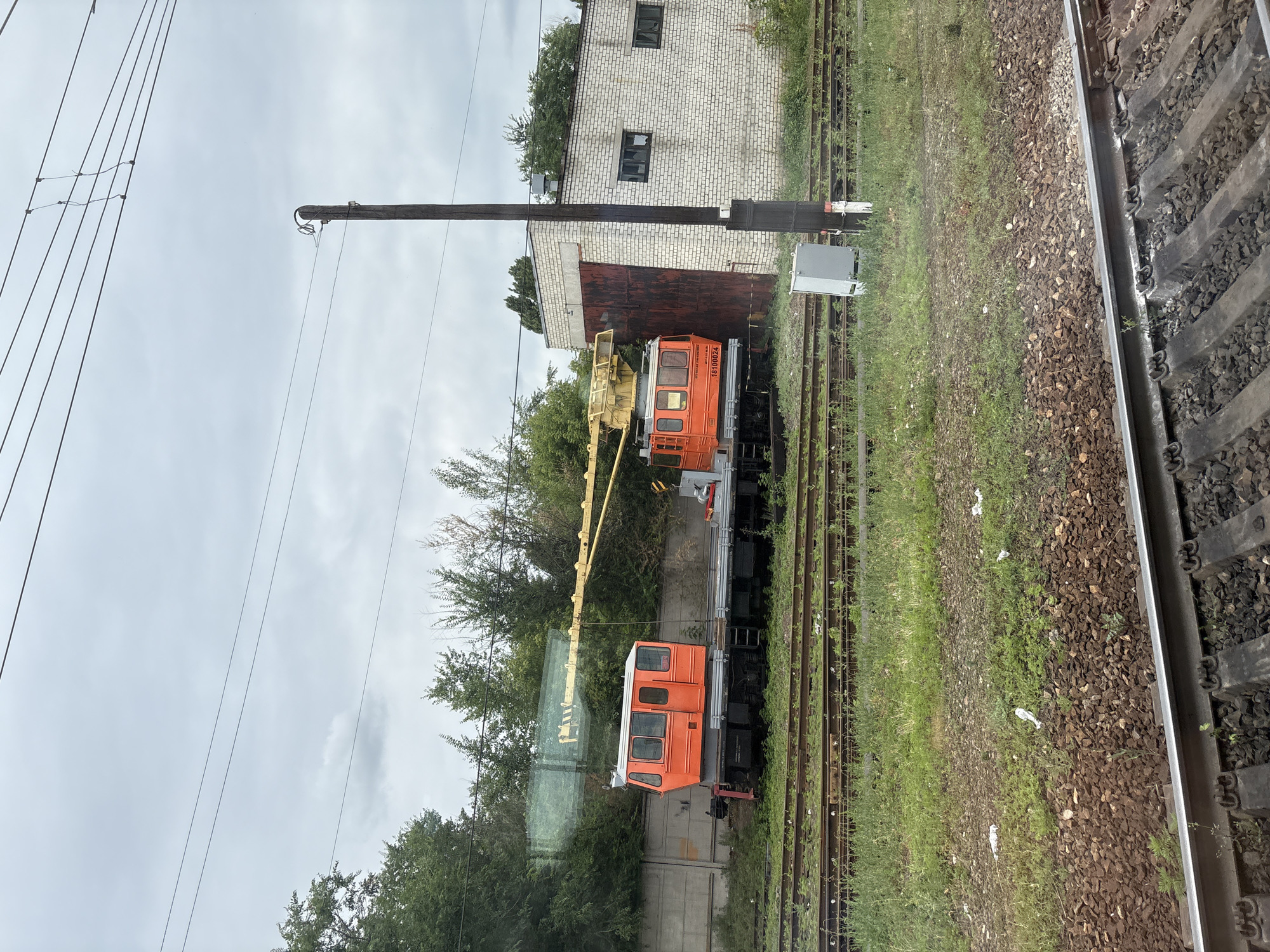 In Povorino, Russia, an outdoor scene depicts a train yard under an overcast sky. Two orange train cars, identified as cranes, are positioned on railway tracks. The crane on the left is closer to the foreground and is partially obscured by trees and a concrete wall. The crane on the right is more visible, featuring a yellow boom and the number "18100024" on its side. Adjacent to the tracks is a white brick building with a large, rusted metal door and two dark-paned windows. A tall, dark utility pole stands near the building. The foreground shows multiple sets of railroad tracks running parallel, with ballast and grass between them. Scattered litter is visible on the ground. Trees and foliage line the background. The overall atmosphere suggests an industrial or maintenance area within the city.
