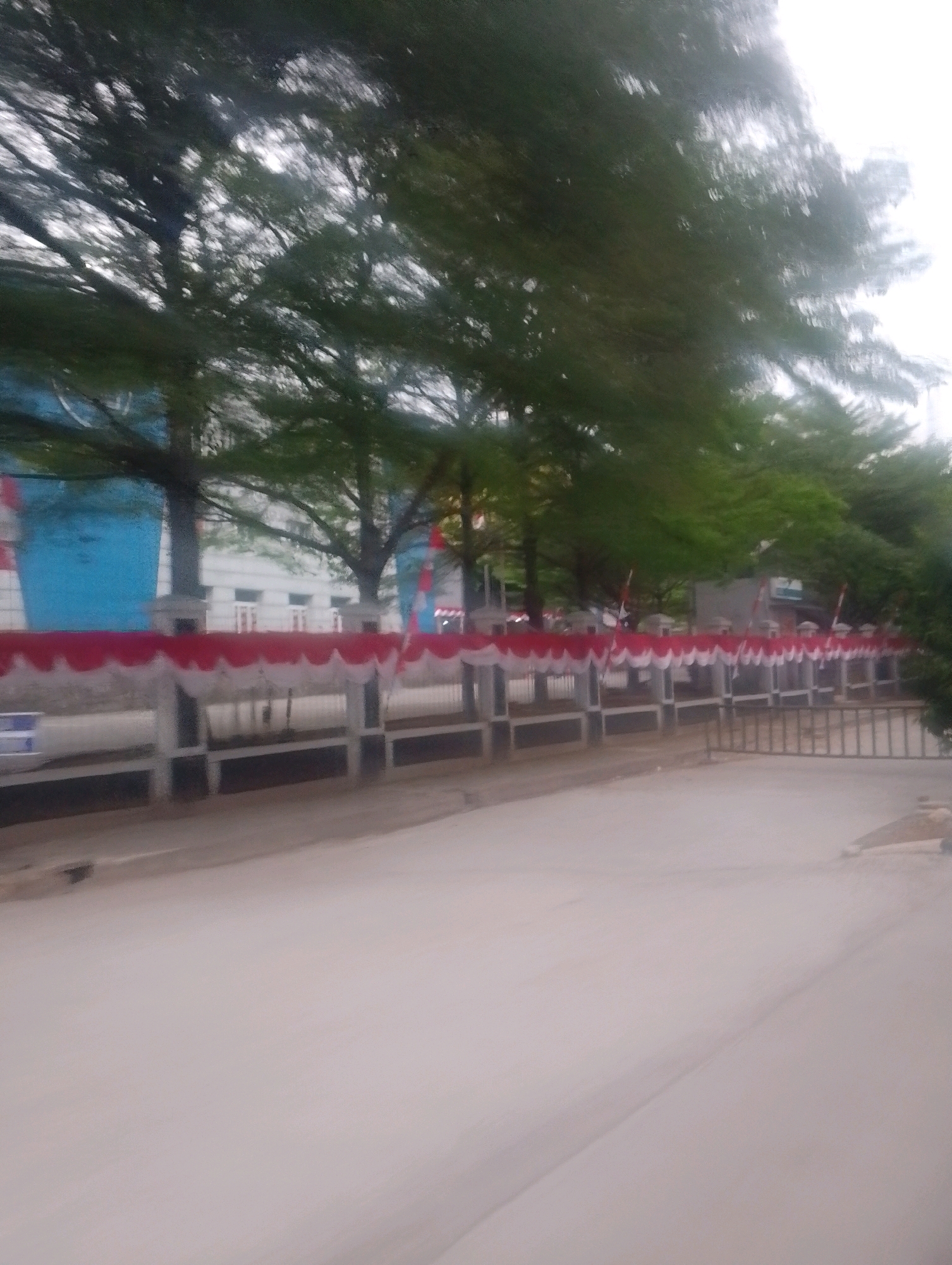 The image depicts a street scene in Depok, Indonesia, with a focus on a building facade adorned with red and white festive banners. The building appears to be a school or government facility, with a large blue cylindrical structure visible to the left. Trees with lush green foliage line the area in front of the building, partially obscuring the view. A fence separates the street from the building's grounds, and a light-colored paved road occupies the foreground. The overall impression is one of a public space decorated for an event.