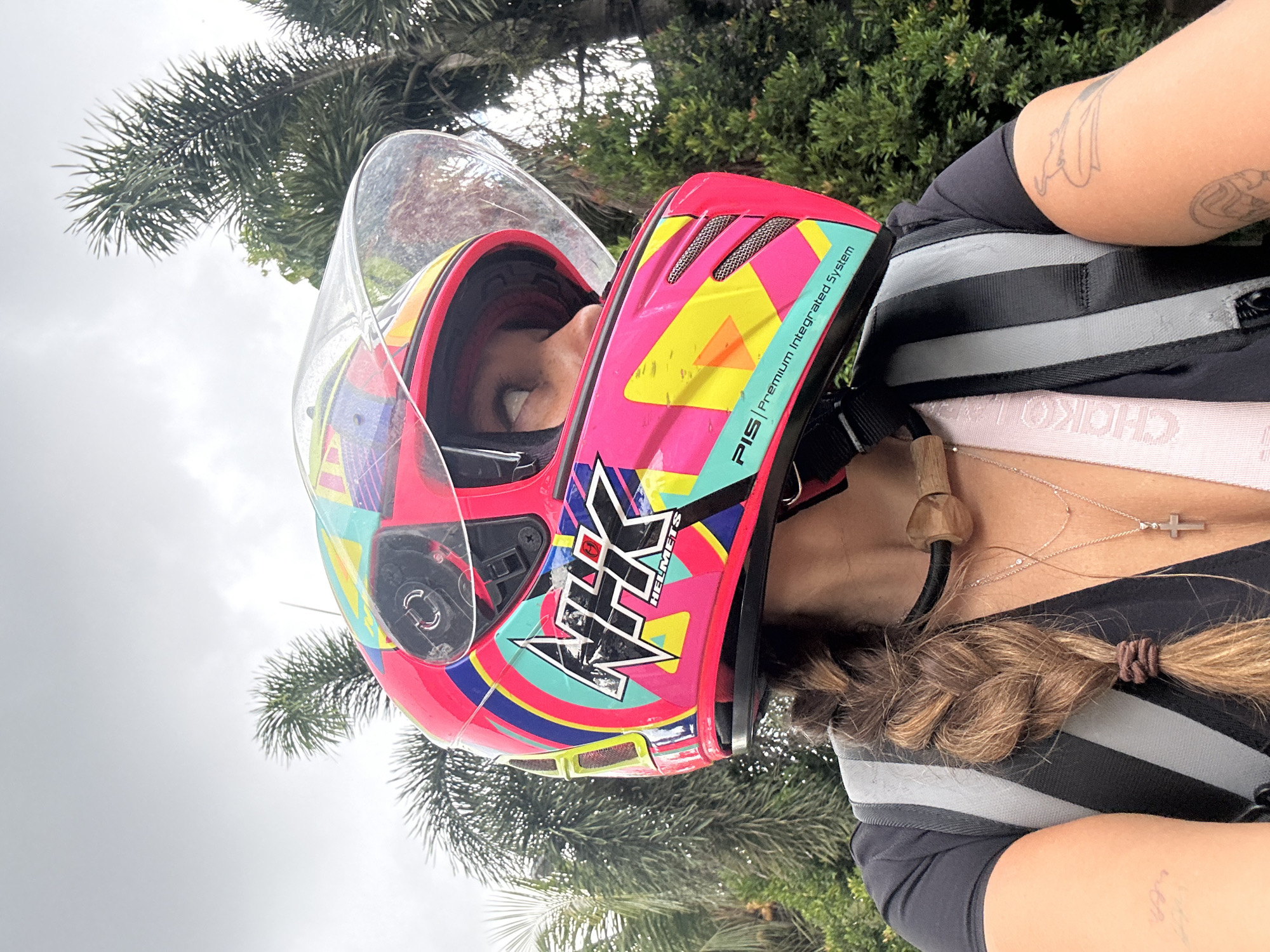 A vertical medium shot shows a woman wearing a multi-colored motorcycle helmet and backpack. The helmet is primarily pink with blue, yellow, and green accents. The helmet is labeled "NHK HELMETS" and "PIS - Premium Integrated System". The woman has a dark braid tied with a brown elastic band on the right. She is wearing a silver necklace with a cross pendant. The backpack is black and grey. The woman's bare arms, adorned with tattoos, are visible on the right. The background is blurred, showing palm trees and a cloudy sky. The location is presumed to be Mengwi, Indonesia, based on implicit visual cues such as the foliage and sky.