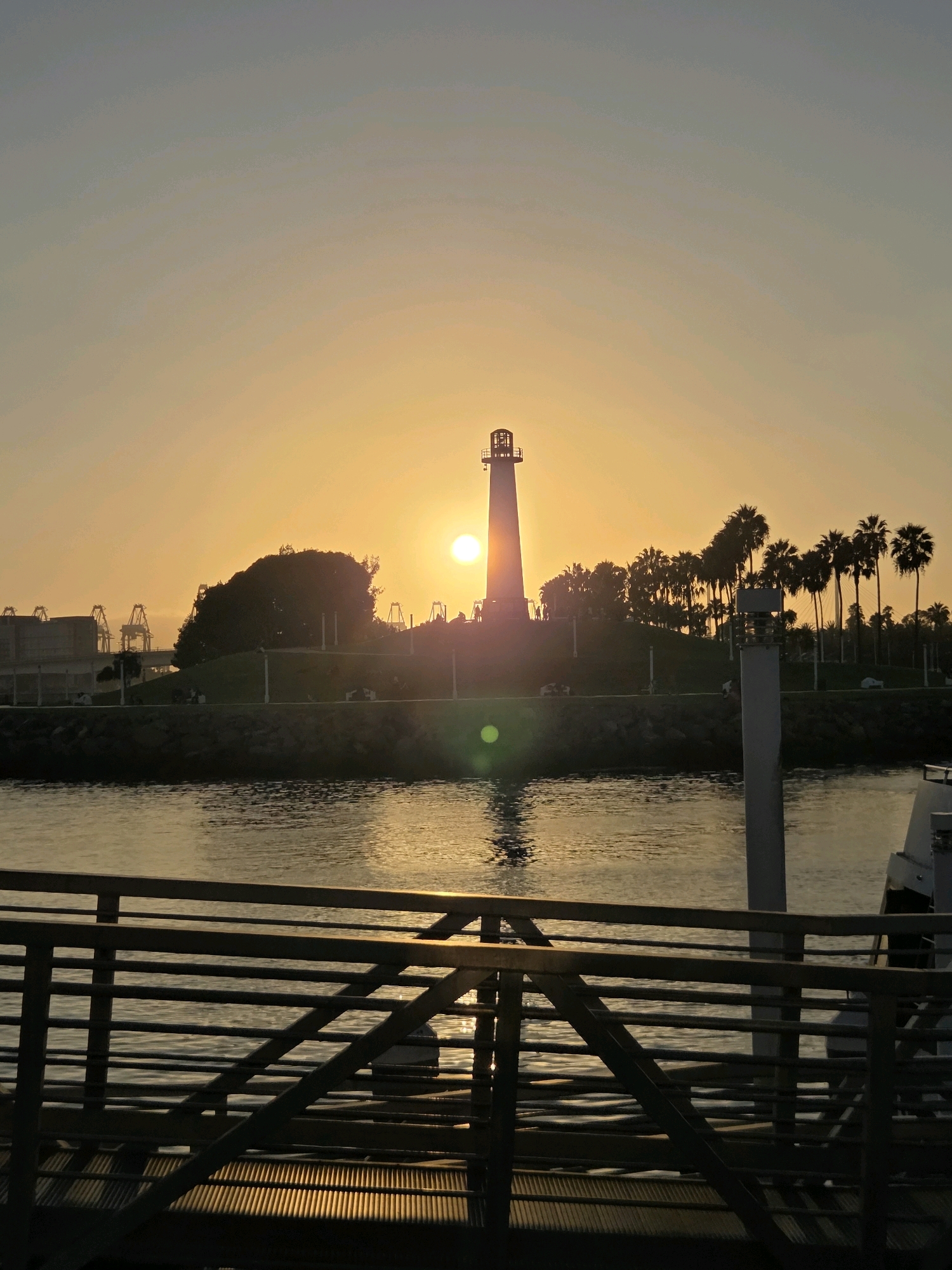 The image depicts a lighthouse silhouetted against a warm, hazy sunset over a body of water. The sky is a gradient of soft oranges and yellows, with the sun prominently positioned near the horizon, just to the right of the lighthouse. The lighthouse itself is tall and white, with a distinct lantern room at its apex. In front of the lighthouse, a line of palm trees stands tall, their fronds catching the fading sunlight. Behind the palm trees, industrial cranes are visible in the distance, suggesting a harbor or port area. The water in the foreground reflects the colors of the sunset, creating a shimmering effect. A dark structure, possibly a pier or boardwalk railing, occupies the bottom portion of the image, with geometric shadows cast across it. The overall scene conveys a sense of tranquility and the end of a day. This scene appears to be the entrance to the Long Beach Harbor, located in Long Beach, California, USA, not Garden Grove, which is an inland city.