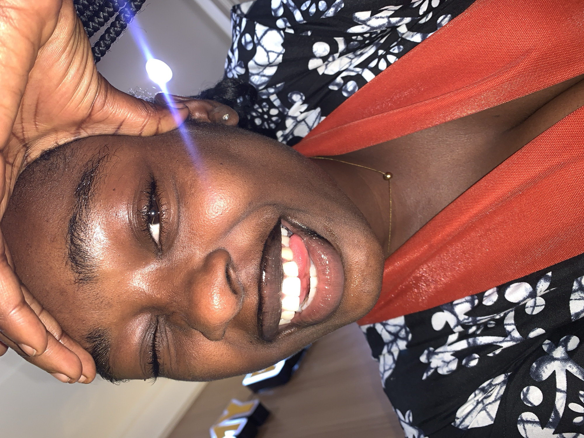 The image is a close-up, selfie-style photograph of a young Black woman. She is looking directly at the camera with her mouth wide open in a smile, her tongue visible. She is winking with her left eye, while her right eye is open. Her right hand is raised and touching her forehead, obscuring part of her hairline. She is wearing a black and white patterned robe or dress, with a bright orange scarf or shawl draped around her neck and chest. A thin gold necklace with a small spherical pendant is visible. A faint blue light glare is present in the upper right quadrant of the image, possibly from a light source or reflection. The background is blurred and appears to be an indoor setting, with hints of what might be signage with yellow lettering. There is no indication of the location being in Gura Top, Nigeria.