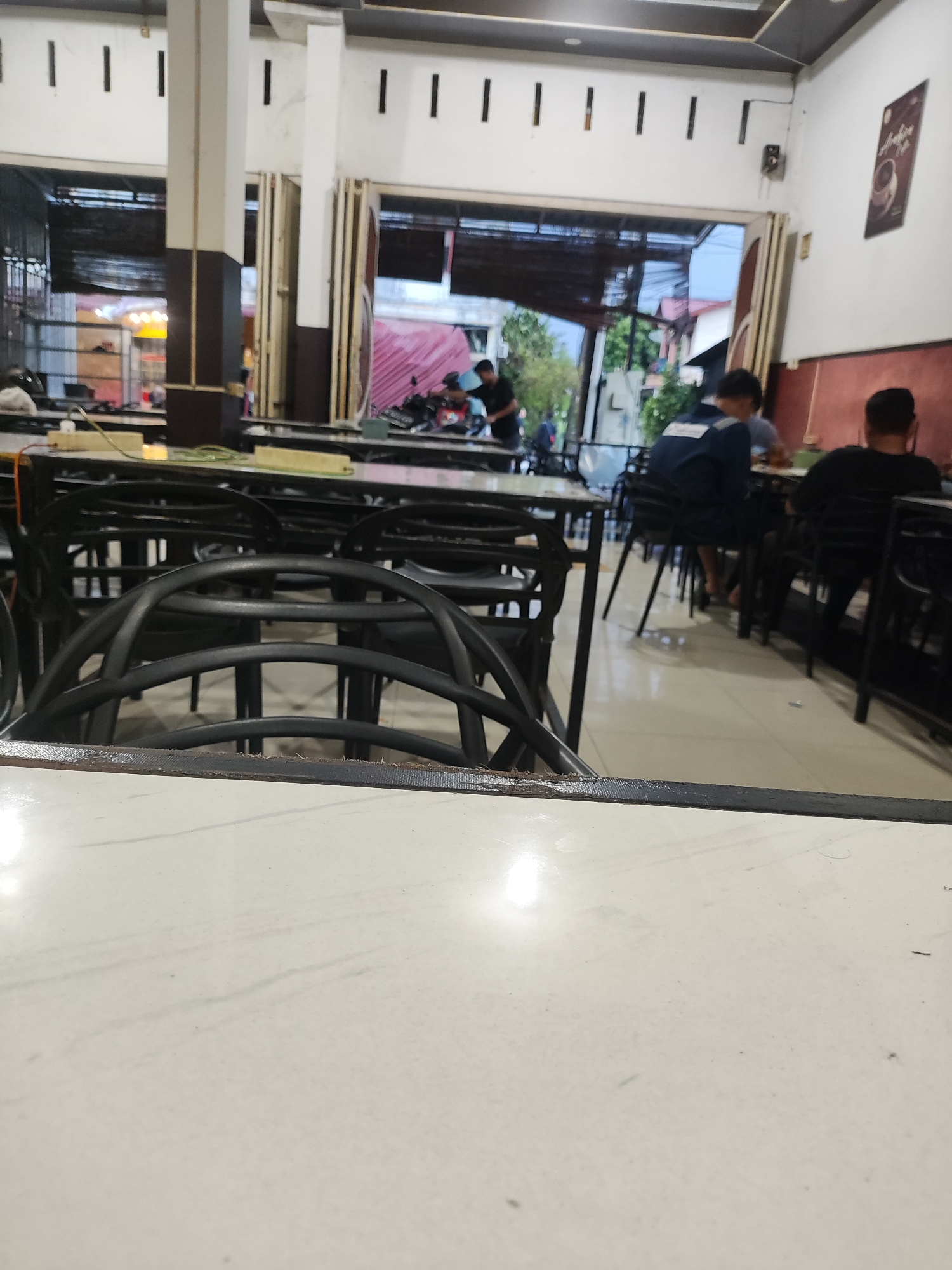 The image shows the interior of a restaurant or cafe in Lhokseumawe, Indonesia. The view is from a low angle, likely a seated position at a table, with a white table top taking up a significant portion of the foreground. A black chair back is also visible in the foreground.
In the midground, there are several tables and chairs, mostly black. People are sitting at some of the tables. A man in a blue shirt is seen from the back, and another man in black is facing away from the camera. Other individuals are visible in the background but are less distinct.
In the background, the open-air facade reveals a street scene with motorbikes parked. The building's architectural features include white pillars with dark bases and a series of narrow vertical windows near the ceiling. A framed poster or sign is visible on the right wall.