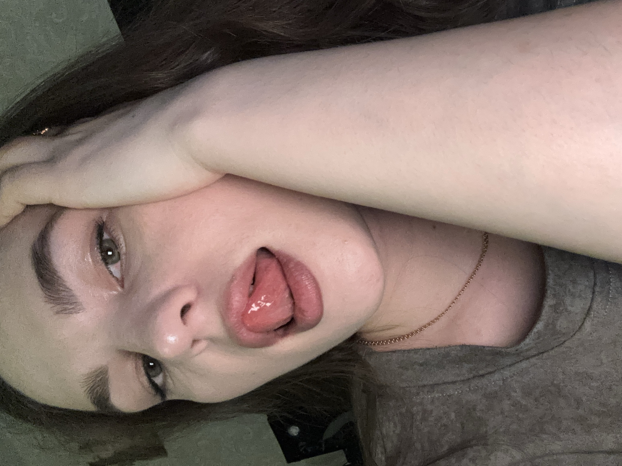 A close-up, angled selfie of a young woman with light skin and brown hair. Her left hand is resting on her forehead. She is wearing a gray t-shirt and a thin gold necklace. Her mouth is open with her tongue sticking out. The background is blurry and dark, suggesting an indoor setting. No specific location in Chaikovsky, Russia can be determined from the image.