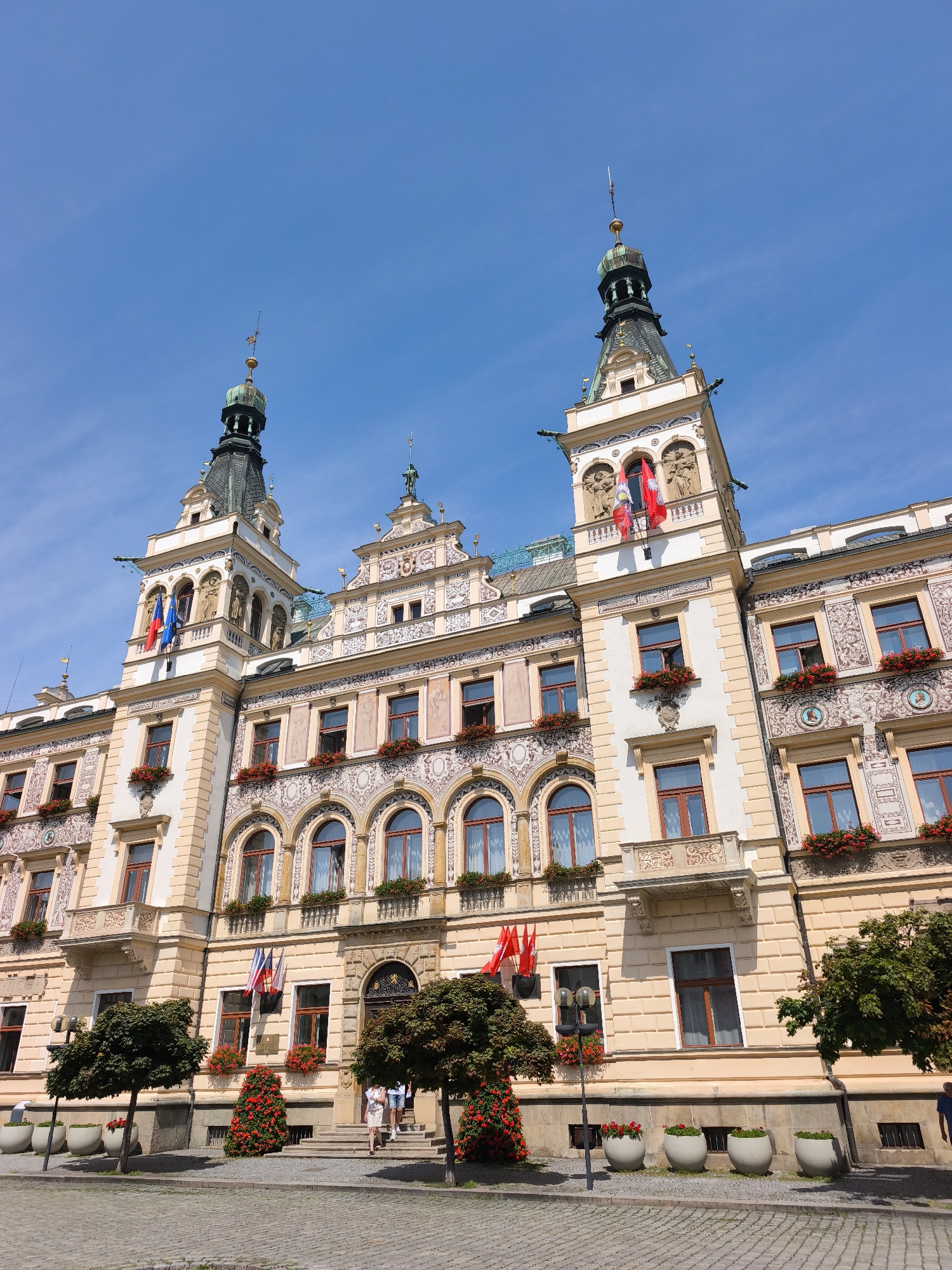 The image shows the Pardubice City Hall, a cream-colored building with ornate decorations, two prominent towers with green spires, and numerous windows. The building is decorated with flags and red flowers in window boxes and planters. Two people are walking up the steps to the entrance of the building. The scene is set on a cobblestone square with trees and planters. The sky is blue with a few wispy clouds.