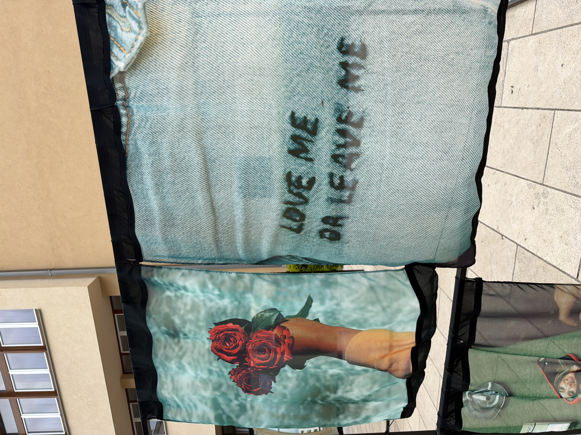 A close-up, eye-level shot captures an outdoor art installation featuring two large fabric panels suspended from a black frame. The panel on the left displays a close-up of a hand holding a bouquet of three red roses against a turquoise, water-like background. The panel on the right, designed to resemble light-wash denim, has the text "LOVE ME OR LEAVE ME" spray-painted in dark lettering. Below these, two smaller fabric panels are visible, one featuring a green background with a circular object and part of a smiling woman's face, and another showing a blurred image. The installation is set against a pale yellow building with brown-trimmed windows and a textured light-brown wall, located on a paved outdoor area. The overall impression is one of a contemporary art display. This scene is observed in Sarajevo, Bosnia and Herzegovina.