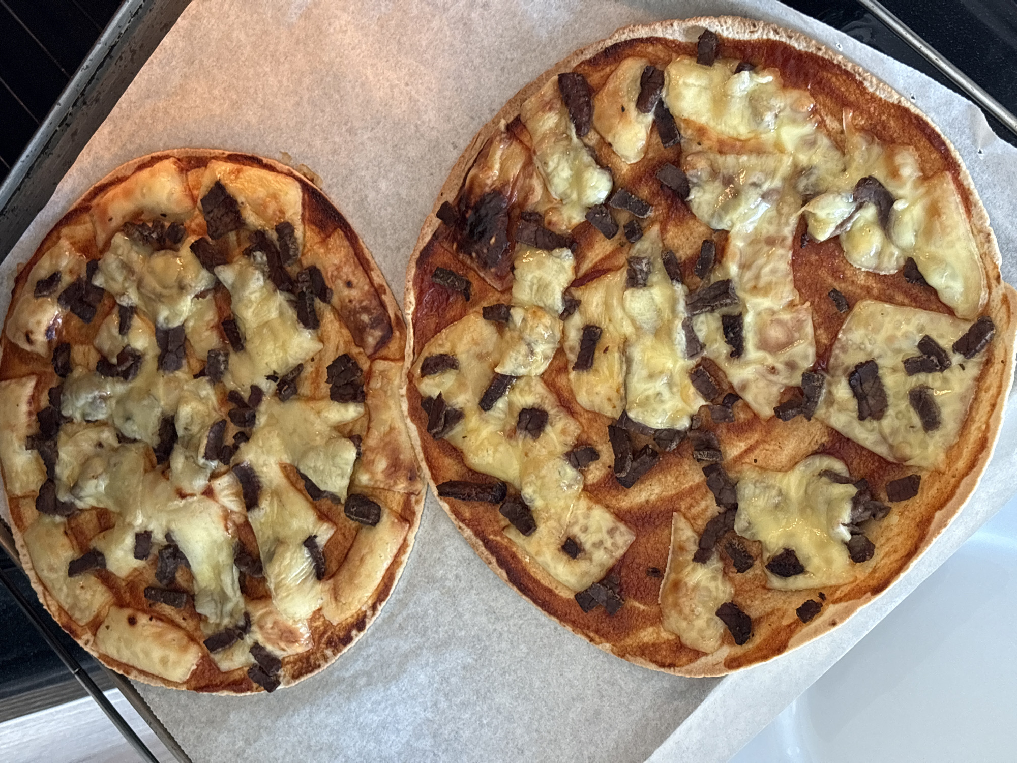 This image captures an overhead view of two freshly baked, small, round flatbreads or pizzas, resting on a sheet of crinkled parchment paper on a dark metal baking tray. The flatbread on the right is larger than the one on the left. Both are generously topped with melted, golden-yellow cheese and numerous small, dark, rectangular pieces, likely cooked meat or a similar topping, distributed across their surfaces. The crusts are golden brown, indicating they are well-baked.
The scene suggests an indoor kitchen setting, with the food items either just removed from an oven or ready to be served. A white surface, possibly a countertop or sink, is partially visible in the bottom right corner of the frame. There are no people visible, and no text can be discerned. While the location context is given as Bar, Montenegro, there are no specific visual cues within the image itself to confirm this geographical location. The indoor lighting does not provide enough information to determine the time of day or the weather outside. The image focuses entirely on the food and its immediate presentation, conveying a sense of homemade preparation.