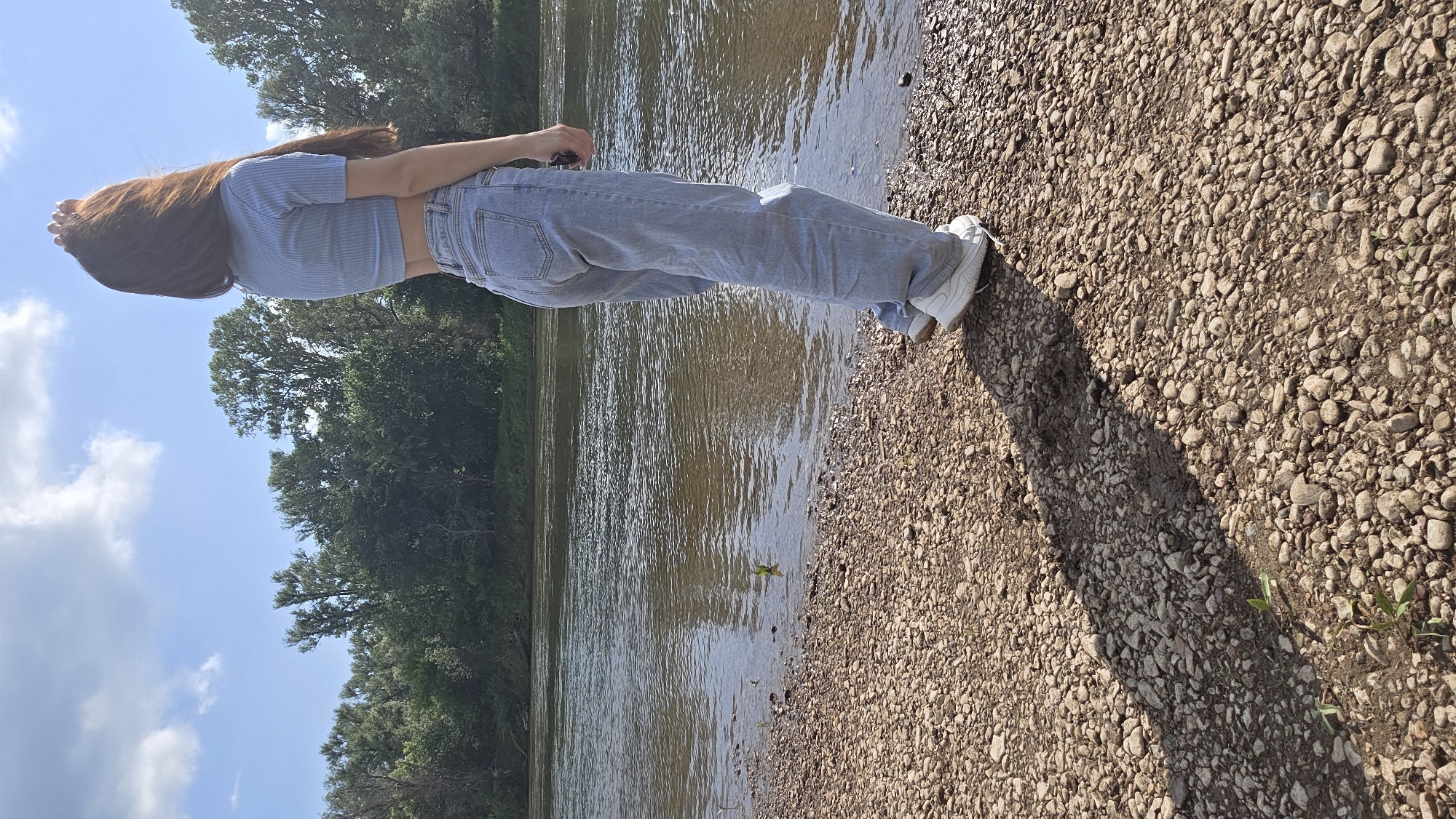 A young woman with long, reddish-brown hair stands on a rocky riverbank, facing away from the camera and looking towards the water. She is wearing a light blue cropped t-shirt and light-wash baggy jeans, along with white sneakers. Her right hand is raised, touching her hair. The river is wide and appears calm, with a gentle ripple on its surface. Across the river, a dense line of green trees forms the opposite bank. The sky above is blue with scattered white clouds. The foreground consists of a textured surface of numerous small, grey and brown stones, with a shadow cast by the woman extending across the ground.