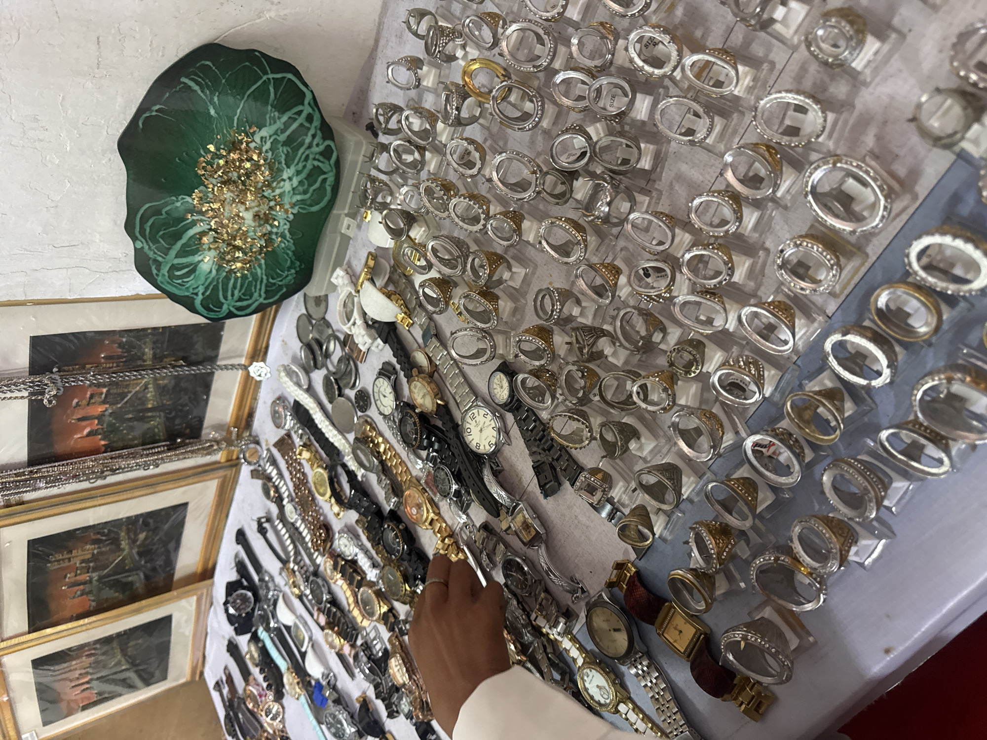 The image depicts a street market stall in Denpasar, Indonesia, displaying a variety of jewelry and watches. The stall features a collection of timepieces, ranging from digital to analog, with various band materials like metal and leather. Alongside the watches, numerous rings are arranged on display stands, most of which appear to be settings without stones, suggesting they are for custom jewelry. A person's hand is visible reaching towards the watches, indicating a potential customer or vendor. In the background, framed artworks, possibly paintings or prints, hang on the wall. A decorative green dish with gold flakes is also prominently displayed. The overall scene suggests a typical marketplace environment focused on accessory sales.