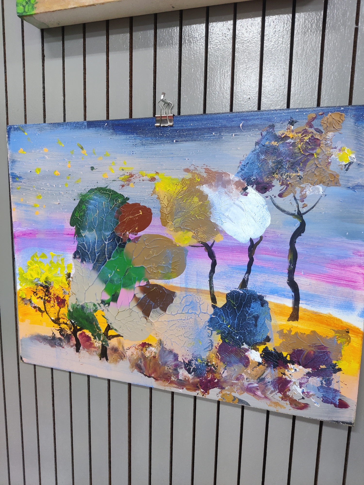 This is a photograph of an abstract painting with a landscape theme, likely depicting trees against a colorful sky. The painting utilizes a thick impasto technique, creating a textured, almost sculptural surface.
The subjects are stylized trees, rendered in bold strokes of various colors. On the left, a tree is painted in shades of green and brown, with a lighter, textured area of white and pink near its base. To the right, two slender trees with dark, twisted trunks stand against a background of layered horizontal strokes in shades of blue, purple, pink, and yellow. The ground beneath the trees is a vibrant yellow, with patches of blue, white, and darker earthy tones. Scattered yellow dots in the sky suggest falling leaves or perhaps a whimsical element.
The painting seems to convey a lively and somewhat chaotic natural scene. The bright, contrasting colors and thick application of paint give the artwork a dynamic and energetic feel. There are no visible people or specific objects, and the scene is purely interpretative.
The setting appears to be outdoors, possibly a field or a stylized forest. The bright colors and horizontal brushstrokes in the sky could suggest a sunset or sunrise, or a vibrant, perhaps even stormy, day. The image is taken from an angle that shows the painting hanging on a wall composed of vertical grey wooden slats. A metal binder clip is visible attaching the top of the painting to the wall.
There is no visible text in the image. The location context of Winterswijk, Netherlands, does not provide any specific cues within the painting itself.