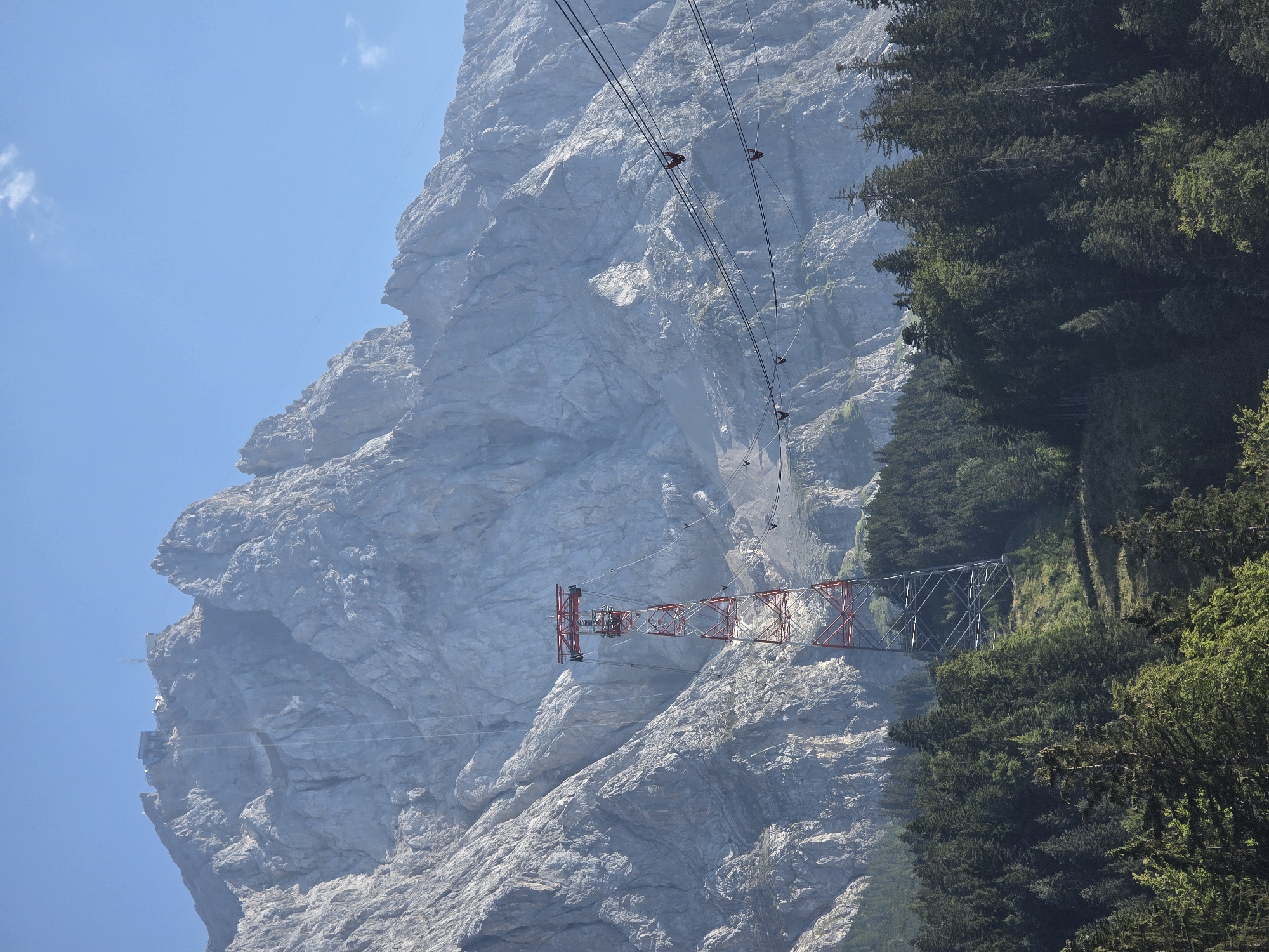 The image captures a majestic mountain range under a clear blue sky, likely in the vicinity of Grainau, Germany, given the presence of the Zugspitze cable car system. In the foreground, a dense forest of evergreen trees blankets the lower slopes of the mountains. Rising from the trees is a striking red and white lattice pylon, a crucial support structure for the cable cars. Several cable cars, small red gondolas, are visible suspended from thick cables, ascending towards the summit. The rugged, rocky face of the mountain dominates the background, showing intricate geological formations and weathered surfaces. A hazy mist softens the distant peaks, adding depth to the landscape. The overall scene depicts a popular tourist destination, highlighting the infrastructure that allows access to the high alpine environment.